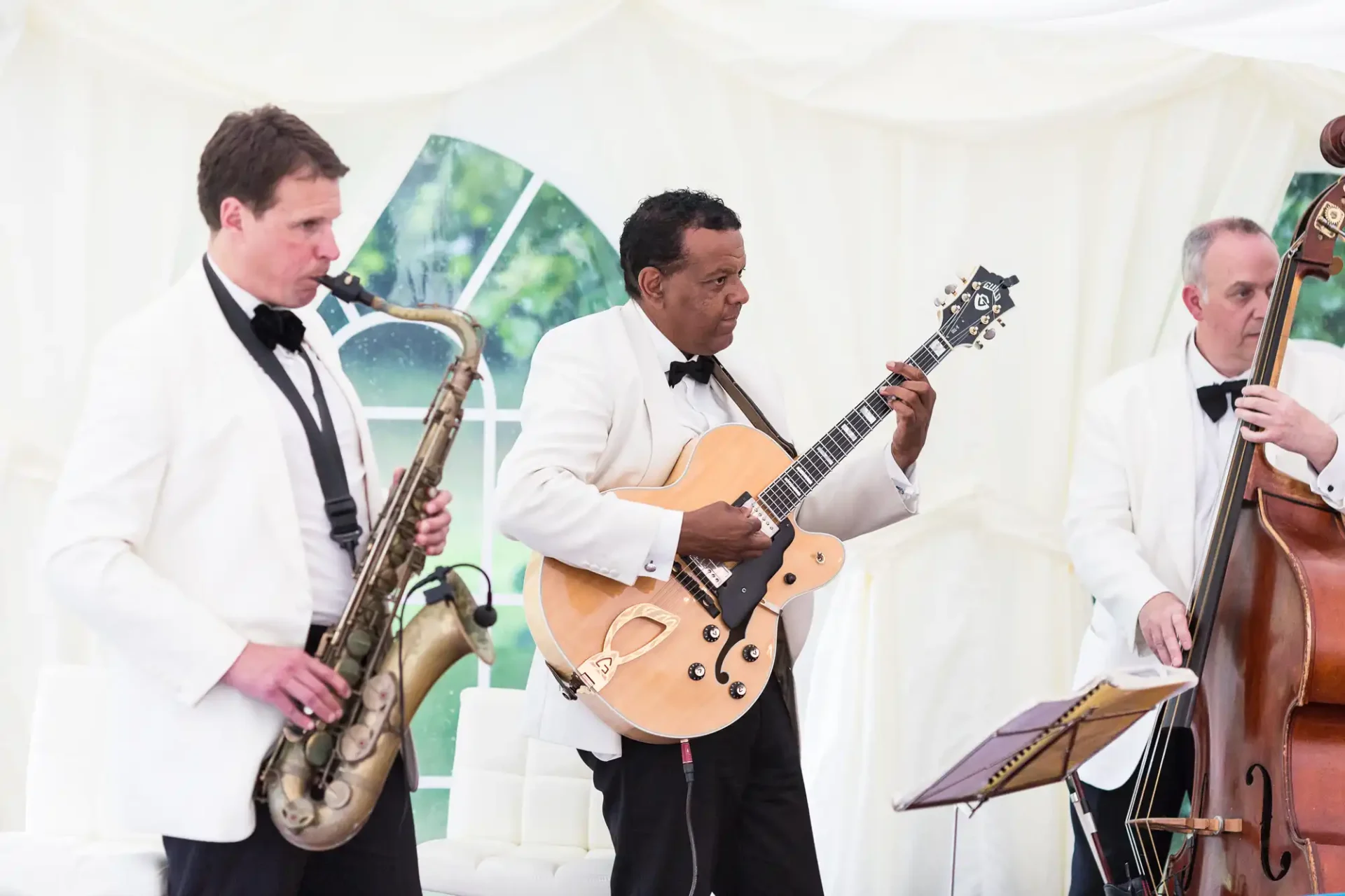 Three musicians in formal attire perform at an event, playing a saxophone, electric guitar, and double bass under a white tent.
