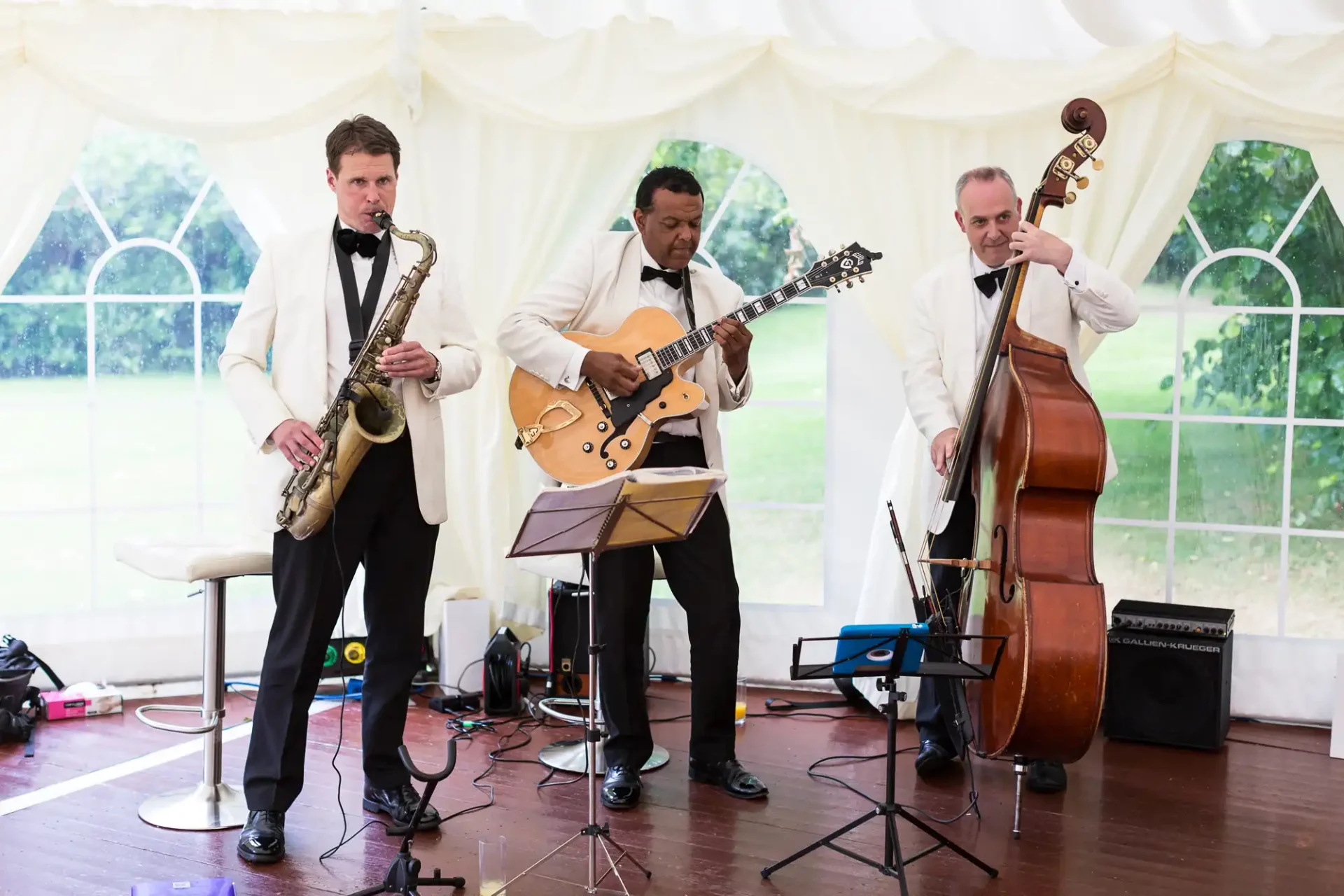 Three musicians perform at an event, playing saxophone, electric guitar, and double bass inside a tent.