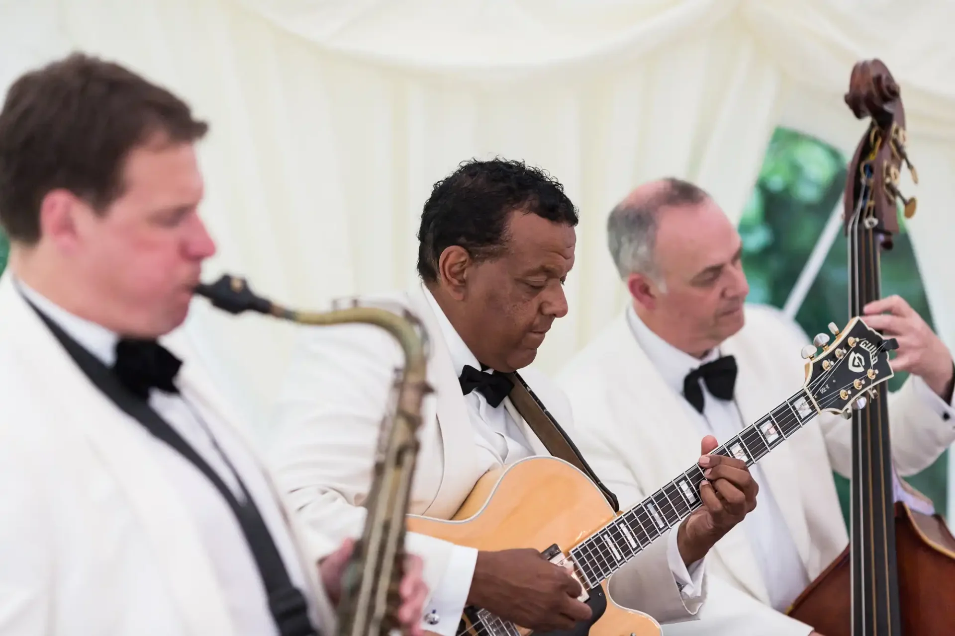 Three musicians in tuxedos playing saxophone, guitar, and double bass at an event inside a marquee tent.