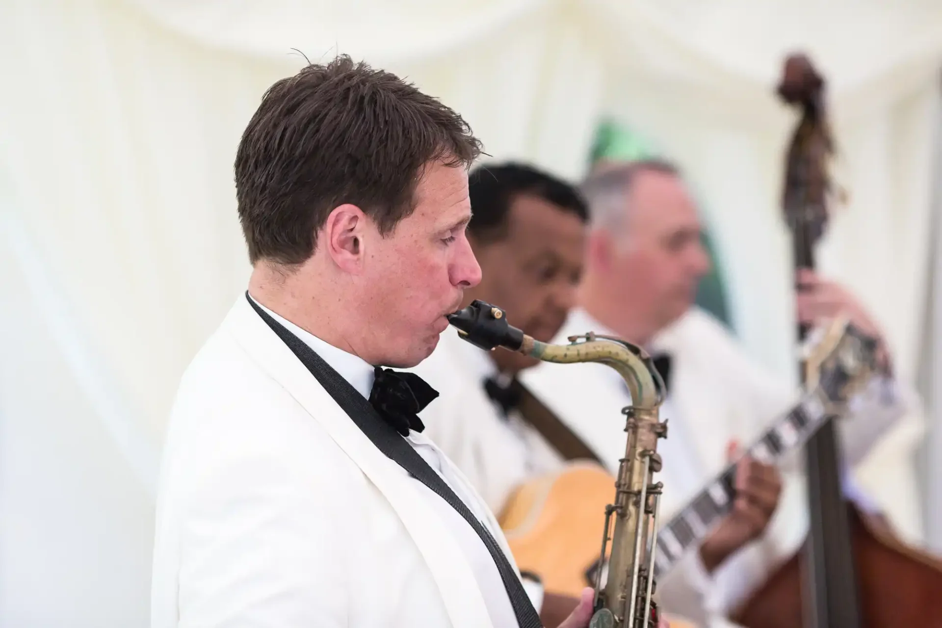 A man in a tuxedo playing the saxophone, accompanied by musicians on guitar and double bass under a white tent.