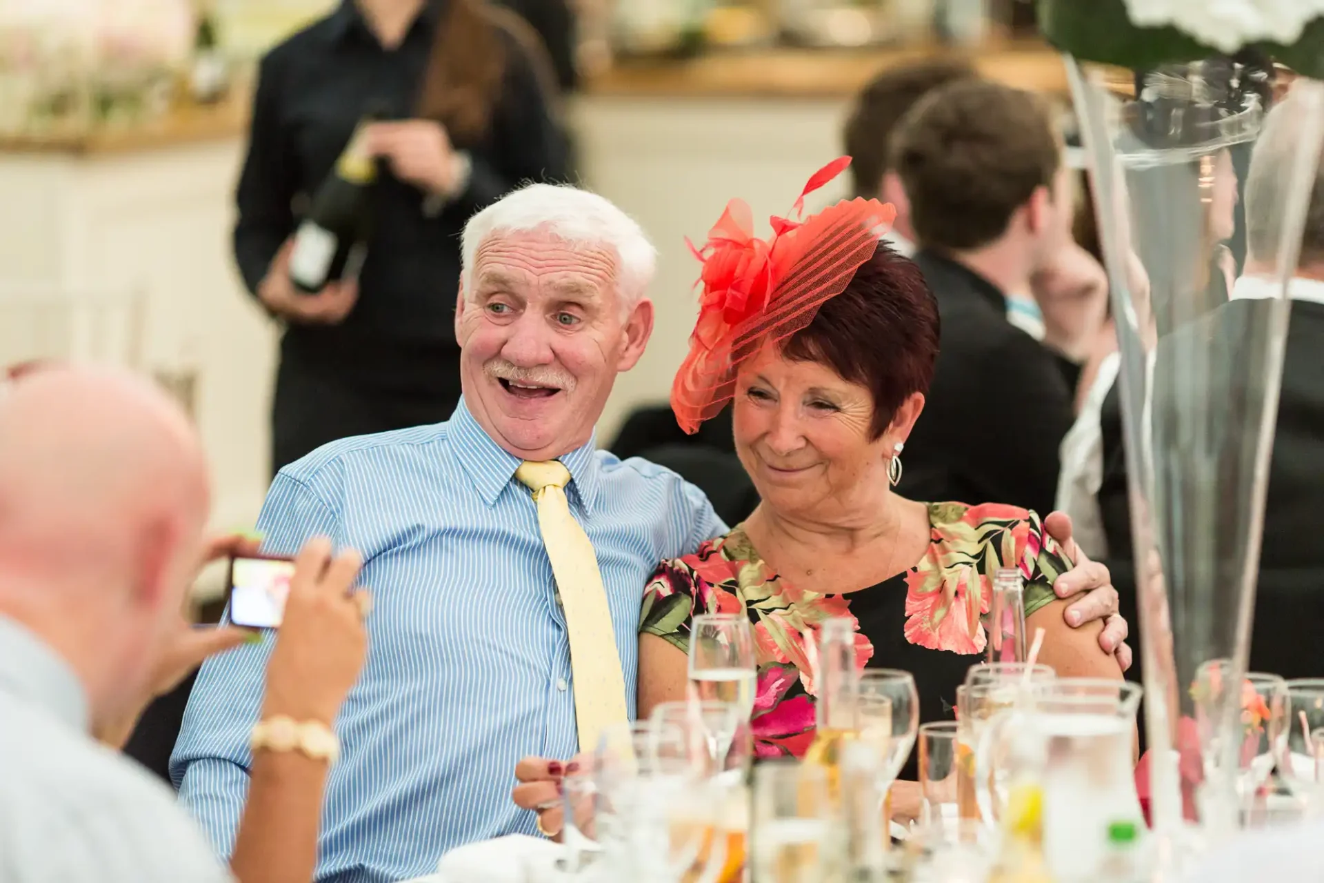 An elderly man in a blue shirt and yellow tie laughs joyously beside a woman in a floral dress and red hat at a formal event.