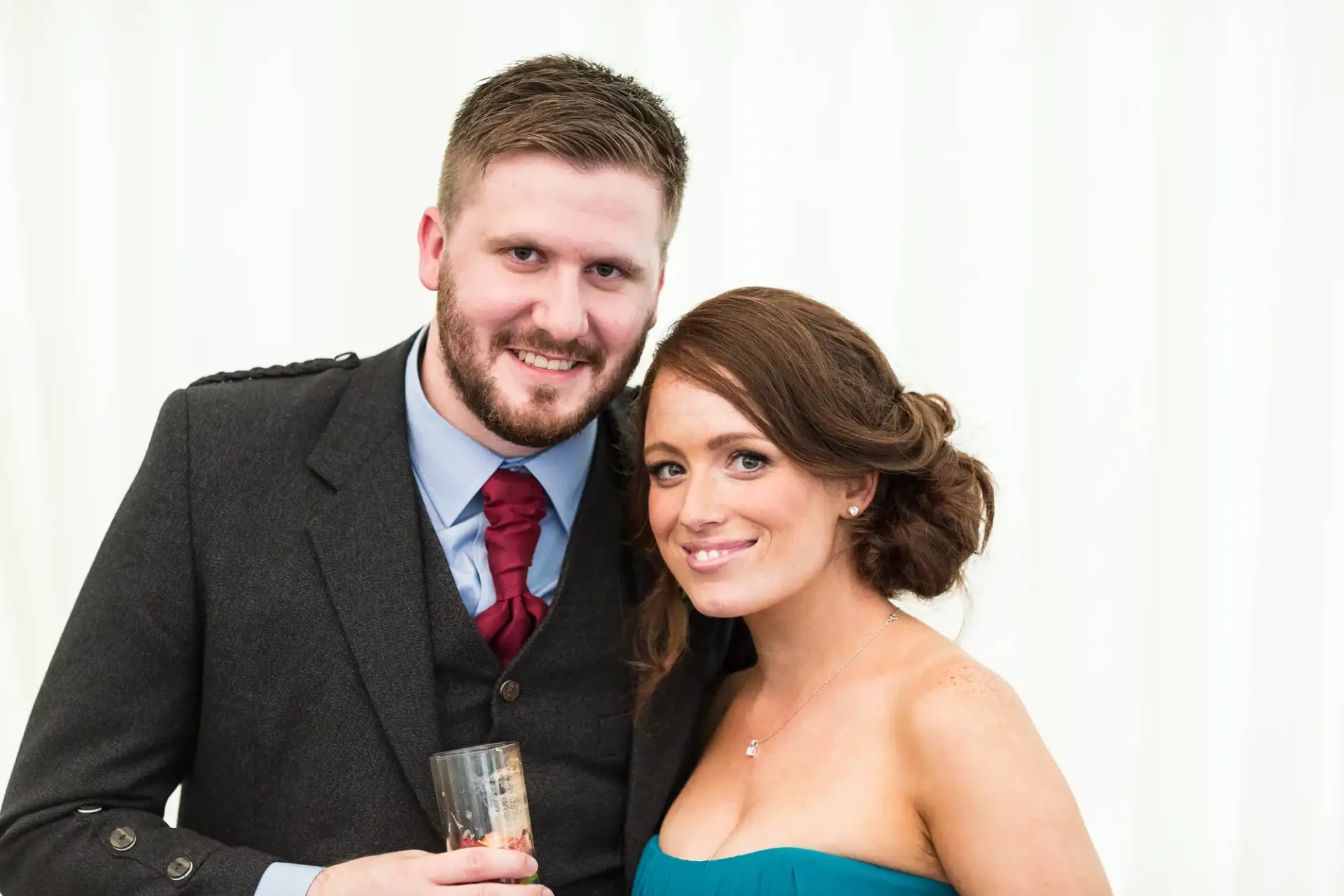 A man and a woman smiling at the camera, the man in a gray suit with a red tie and the woman in a teal dress, both holding a glass.