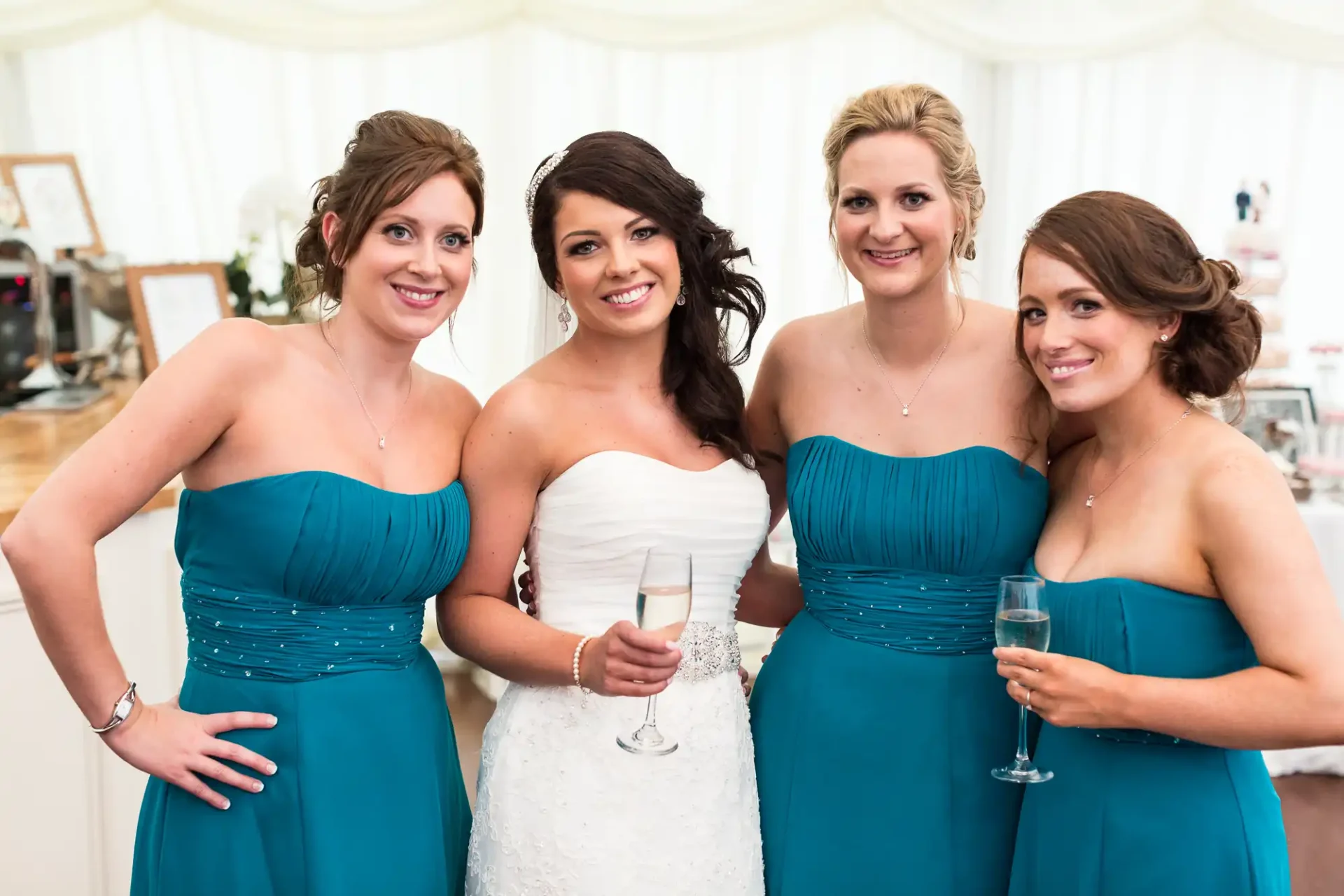 A bride in a white dress and three bridesmaids in teal dresses smiling inside a wedding tent, two holding champagne glasses.