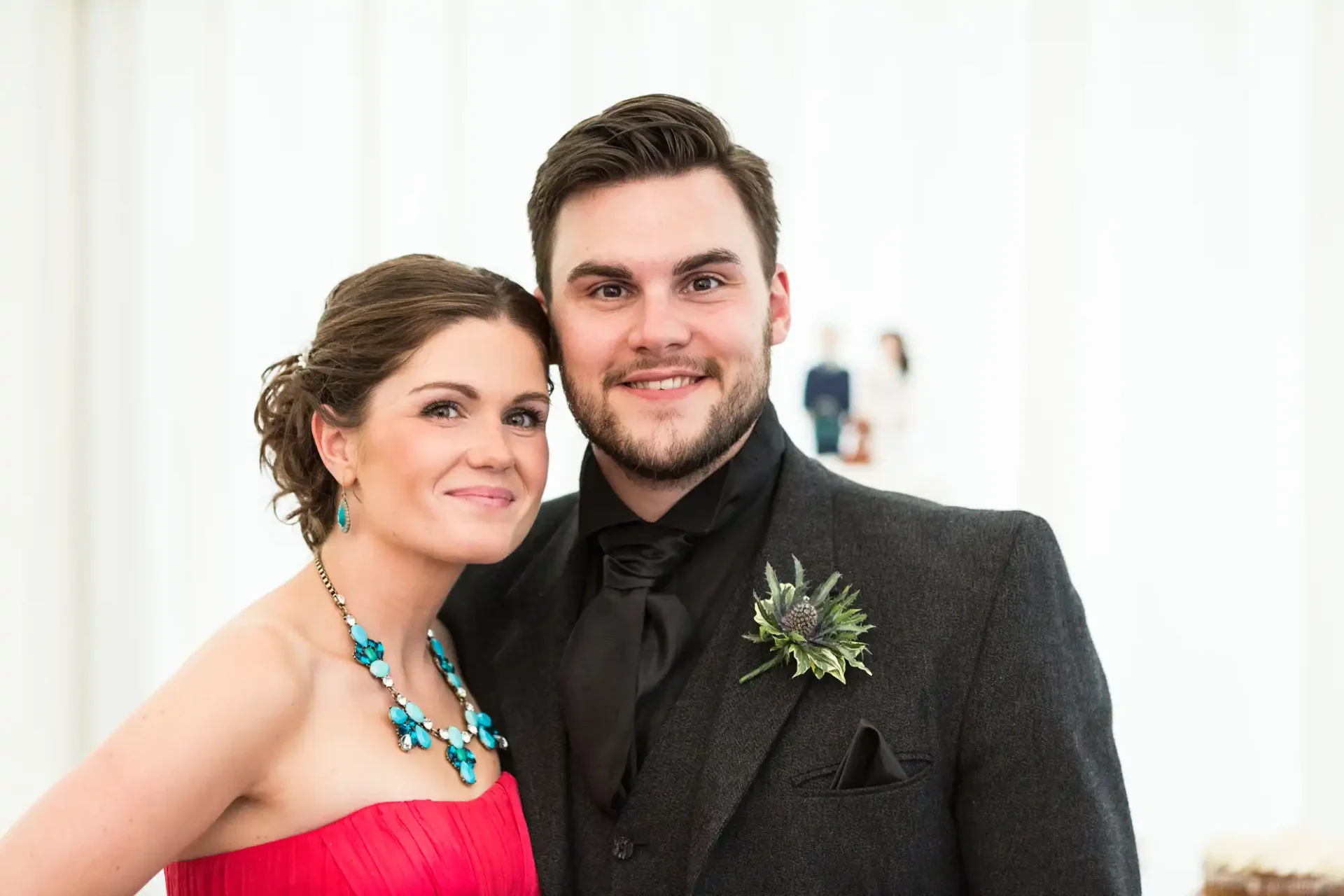 A couple dressed in formal attire smiling at a wedding, with the woman in a red dress and blue necklace, and the man in a gray suit with a black bow tie.