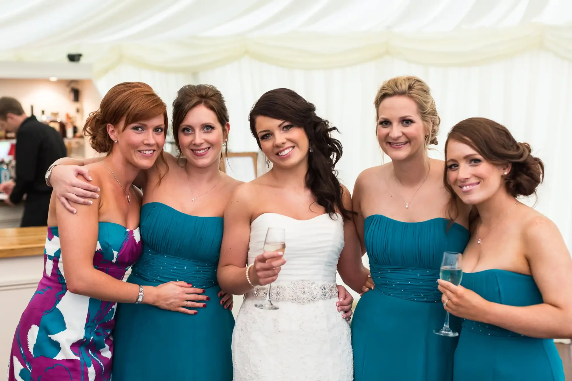 Five women at a wedding reception, smiling for the camera, four in matching blue dresses and the bride in white, holding champagne glasses.