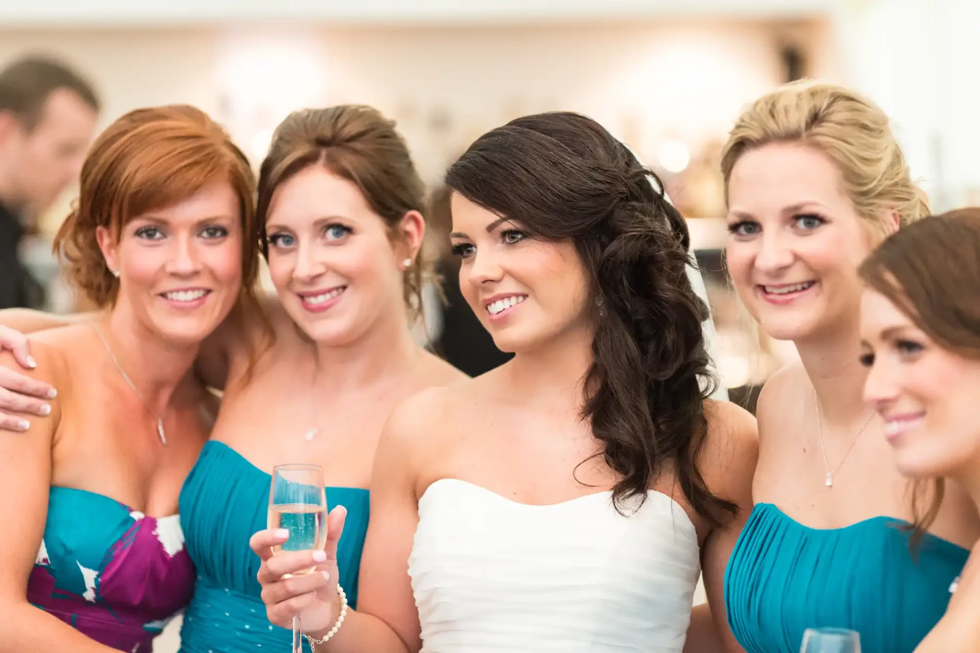 Five women in formal attire at an event, with one in a white dress holding a champagne flute, surrounded by others in matching blue dresses.