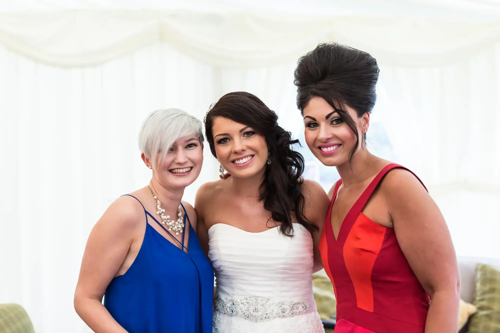 Three women smiling at a wedding, one in a white bridal gown and the others in blue and red dresses, inside a white tent.
