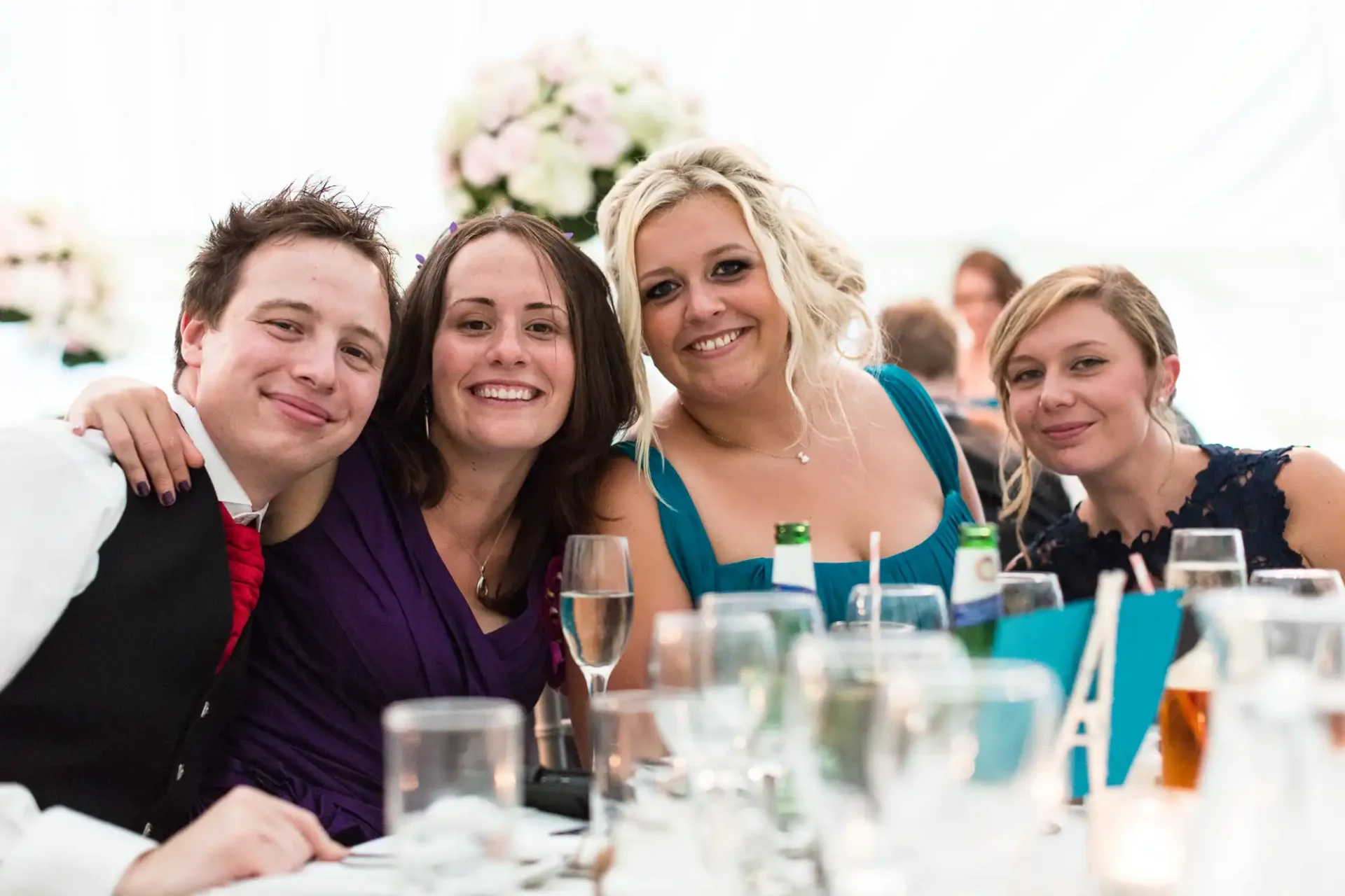 Four friends smiling at a wedding reception table, surrounded by drinks and floral decorations.