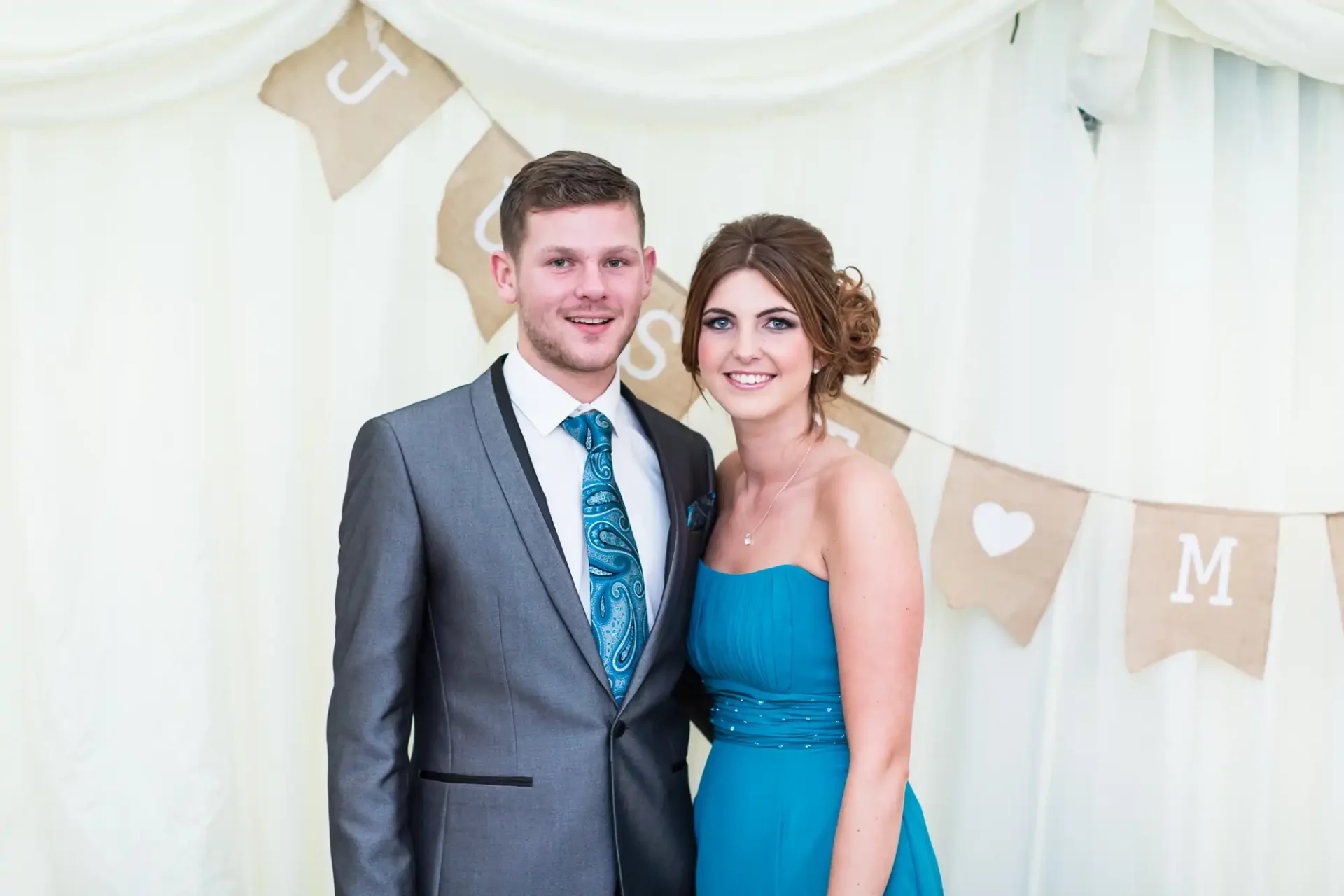 A couple in formal attire smiling for a photo at a wedding, with decorative bunting in the background.