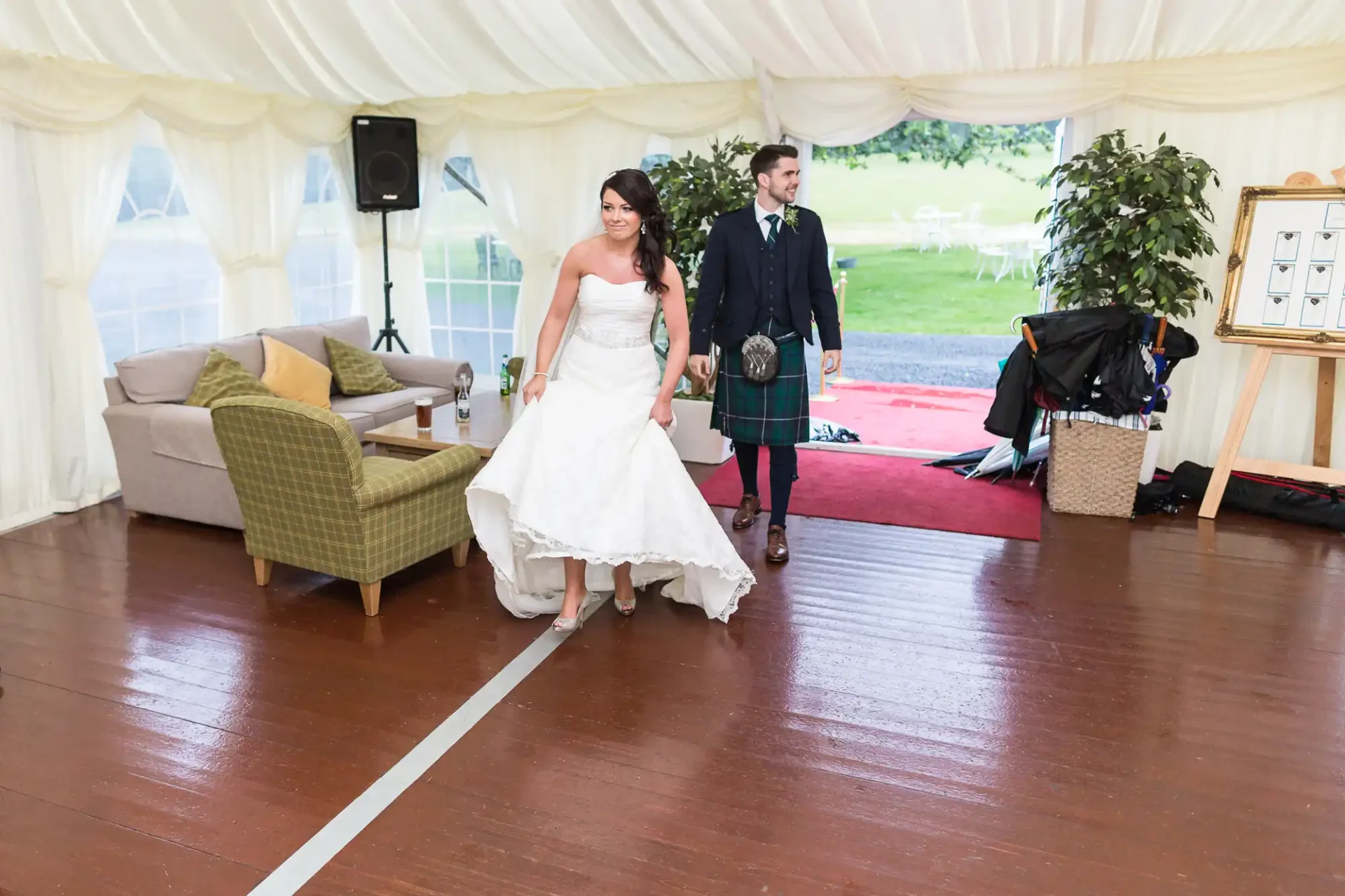 A bride and groom walking hand in hand inside a tent at a wedding reception, with furniture and a green outdoor setting visible in the background.