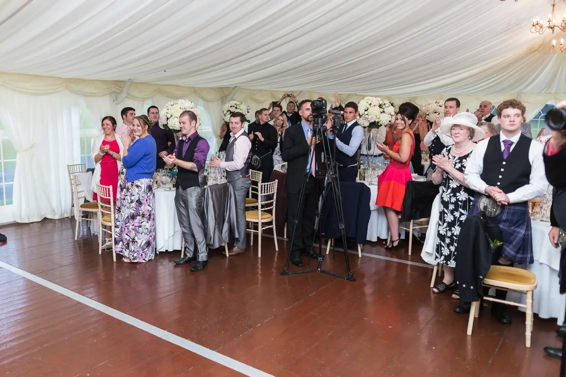 Guests at a wedding reception standing and watching an event inside a marquee, some holding cameras.