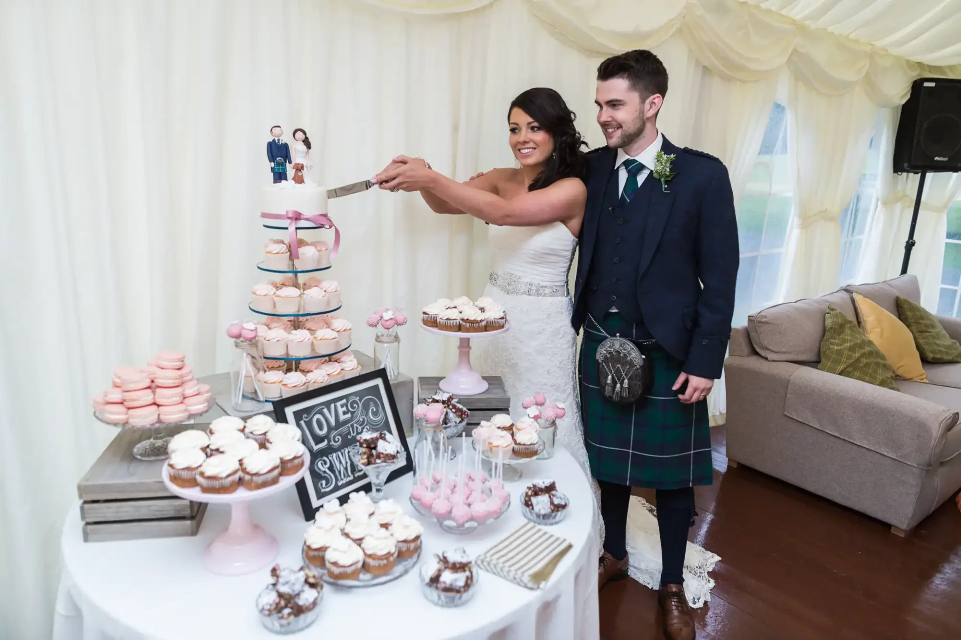 A bride and groom in wedding attire cutting a cake at a dessert table filled with cupcakes and sweets.