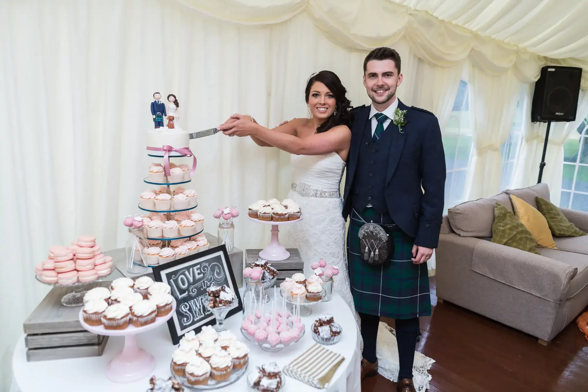 A bride in a white dress and a groom in a kilt cutting a wedding cake, surrounded by cupcakes and desserts in a decorated tent.