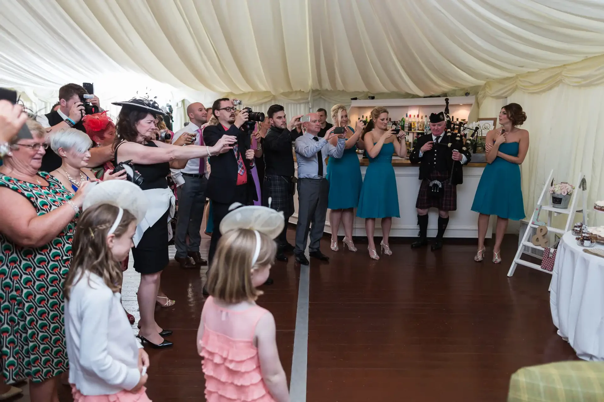 Guests in formal attire taking photos of a bride and groom entering a wedding reception tent, with a bagpiper present.