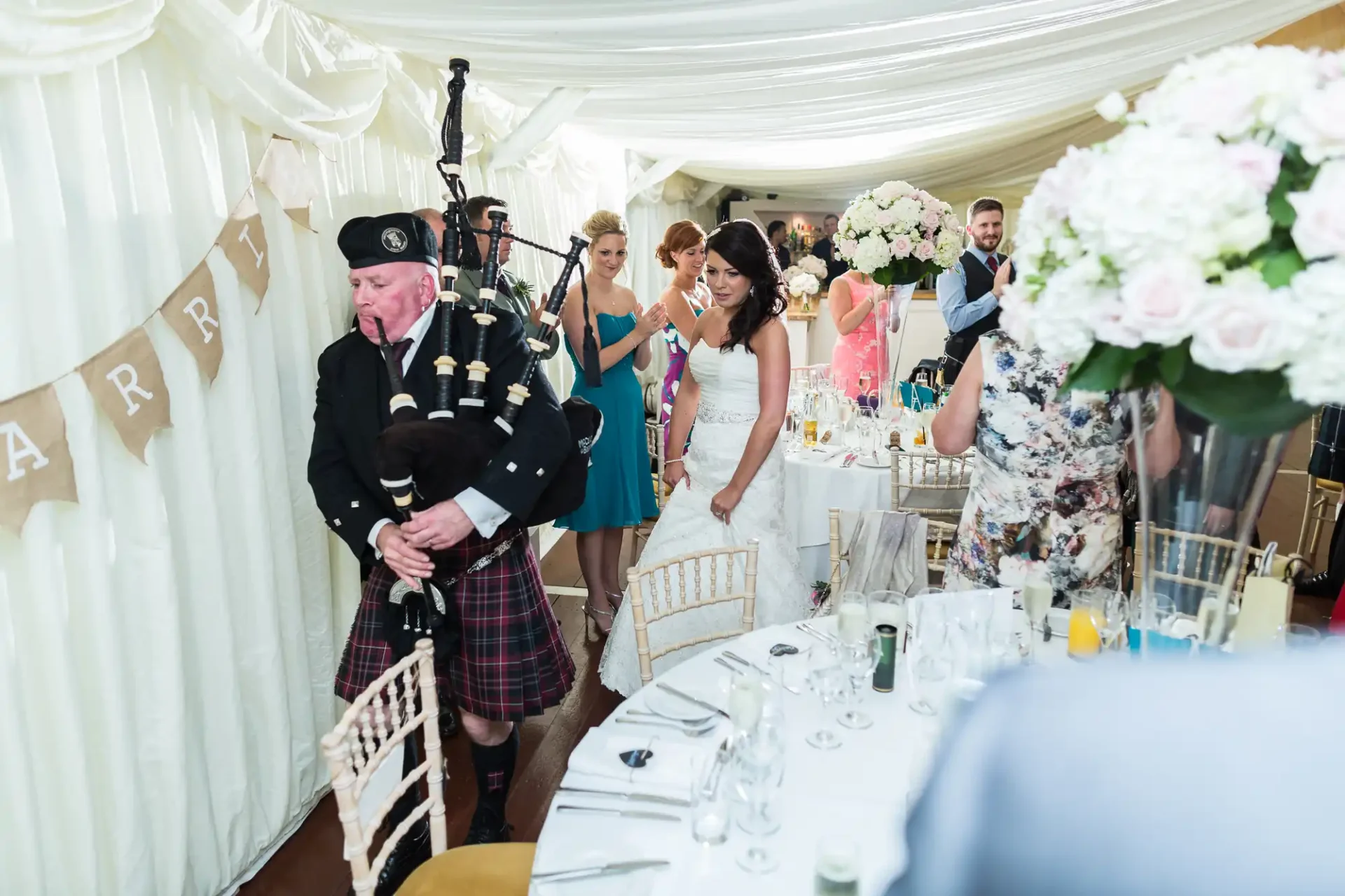 A bagpiper leads a bride through a wedding reception tent, with guests standing and applauding.