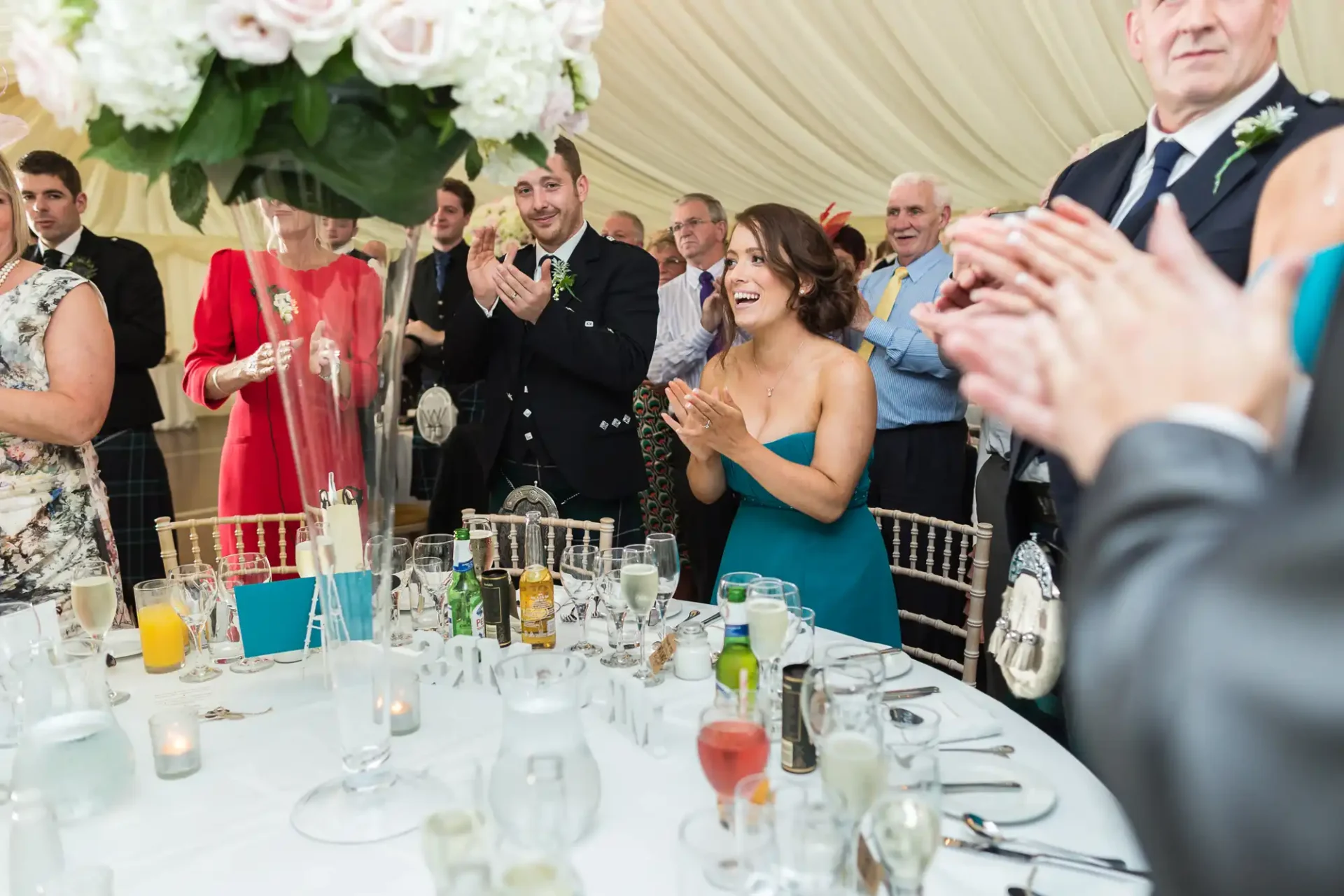 Wedding guests clapping and laughing around a festively decorated table inside a tent.