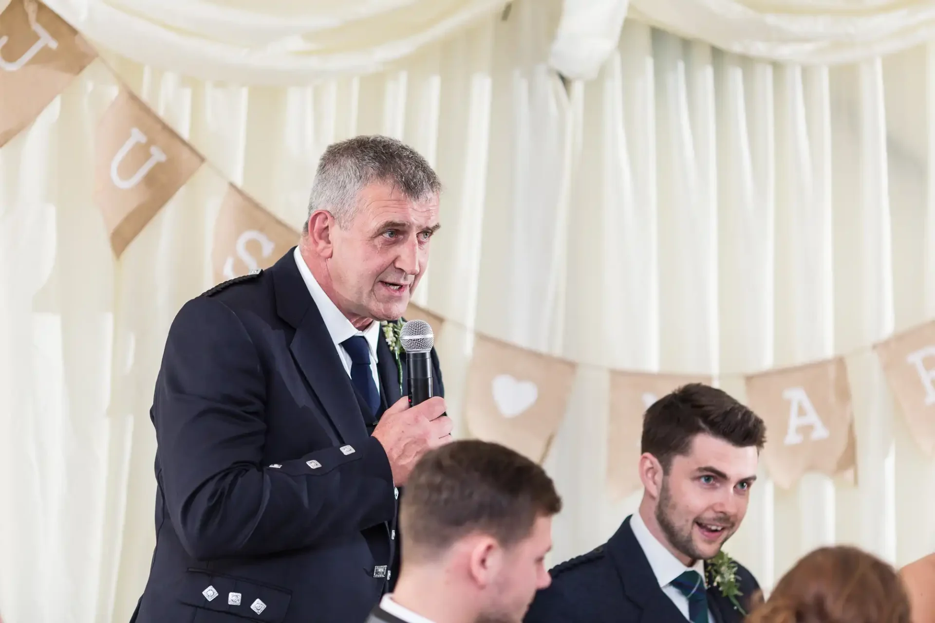 An older man in a suit speaks into a microphone at a wedding reception, with a banner reading "just married" in the background and a young man looking on.