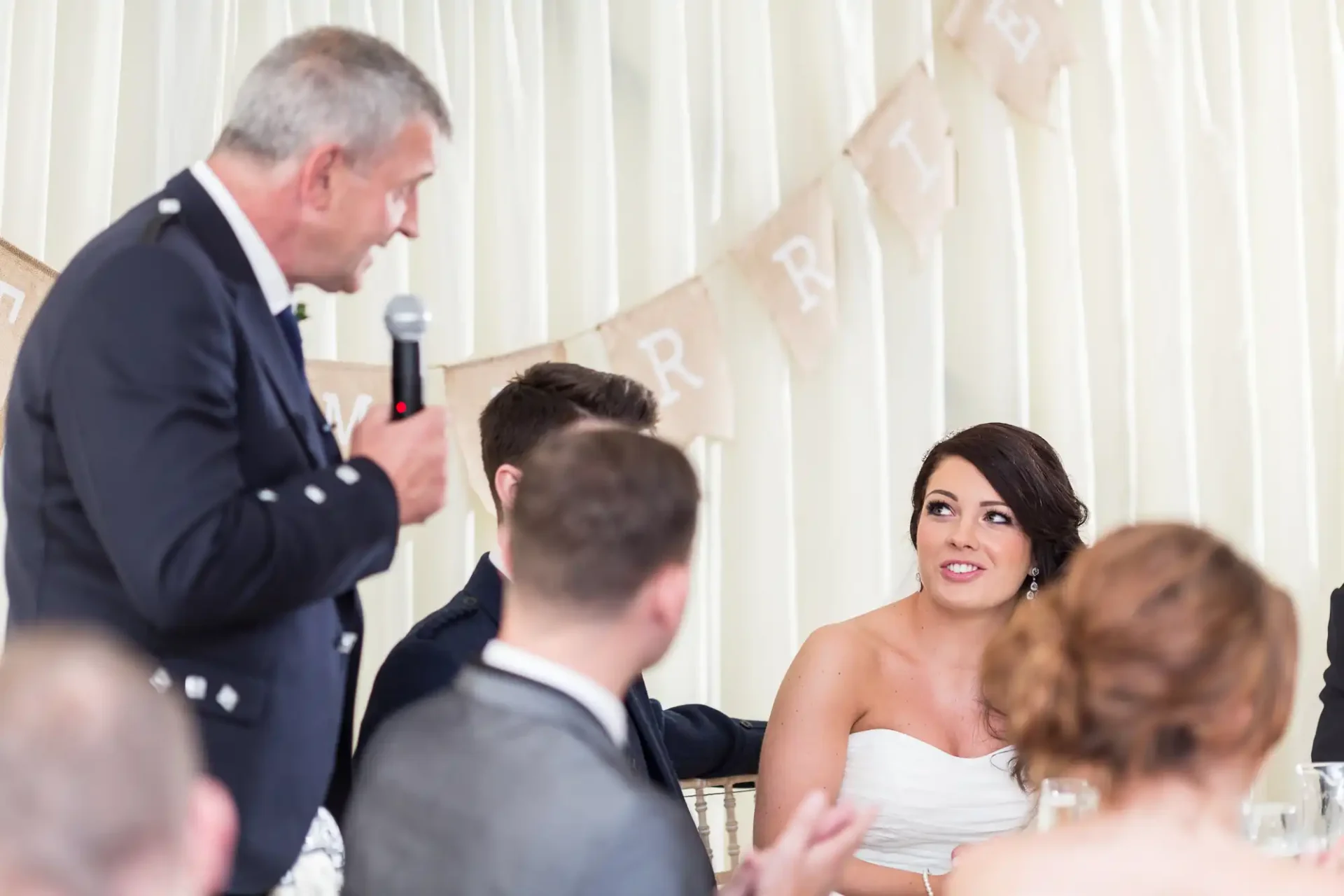 A bride attentively listening to a man giving a speech at a wedding reception, with guests seated around the table.