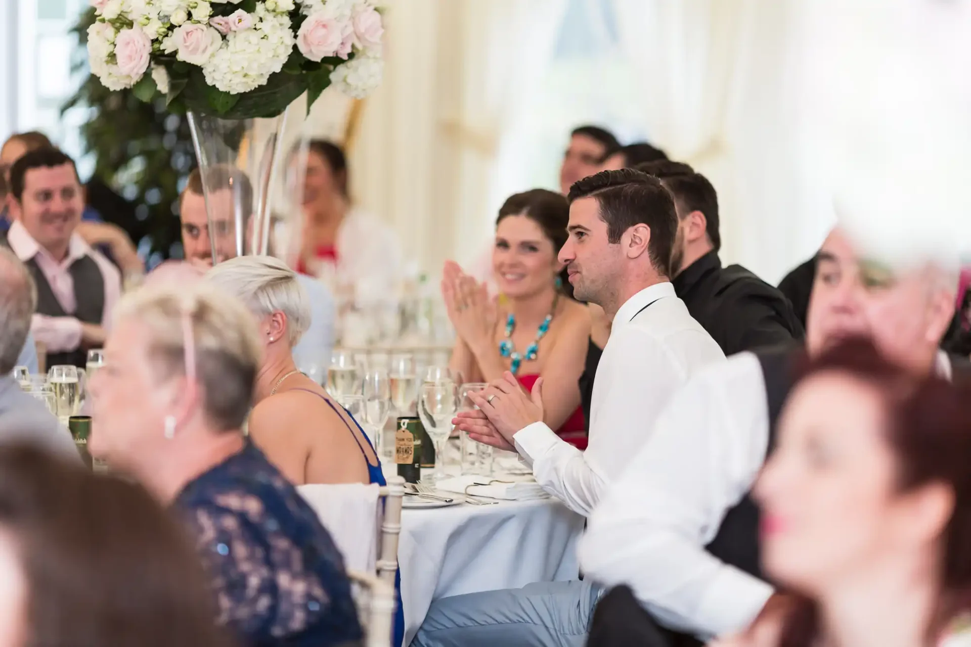 Guests seated at tables, enjoying a conversation during a formal event, with focus on a man in a white shirt.