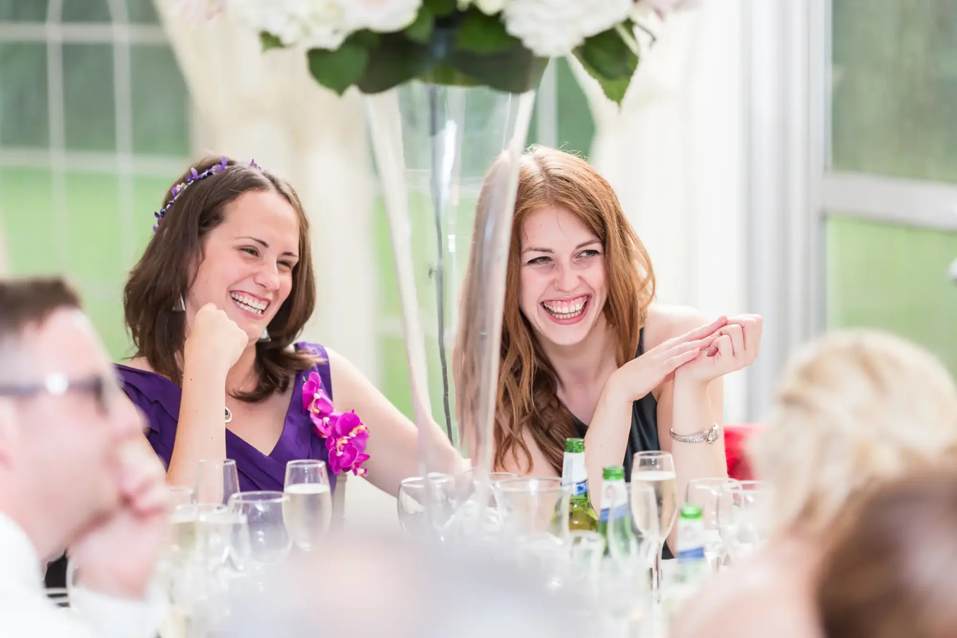 Two women laughing joyfully at a wedding reception table, surrounded by guests and floral decorations.