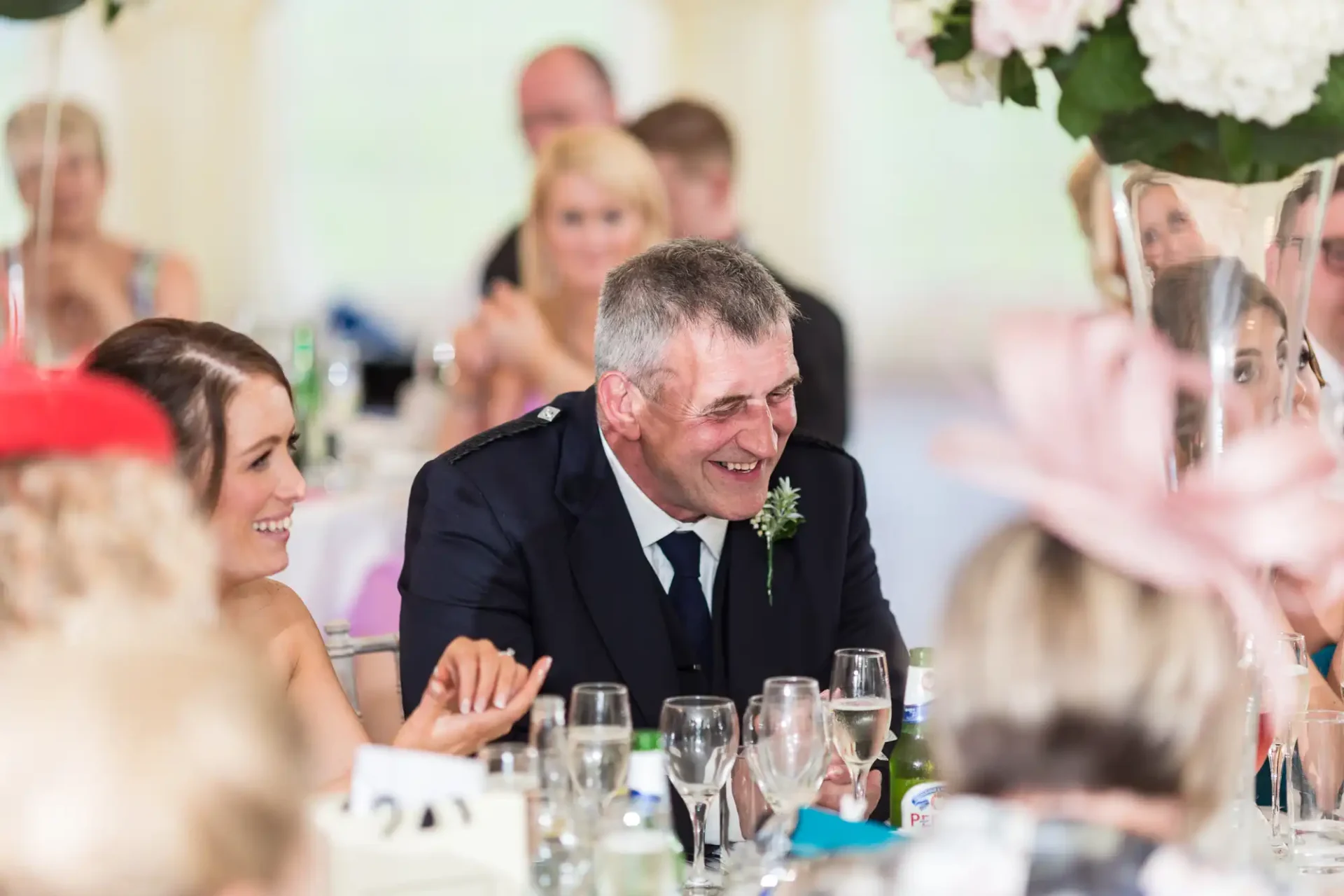 A man in a suit laughing at a decorated table during a wedding reception, surrounded by other guests.