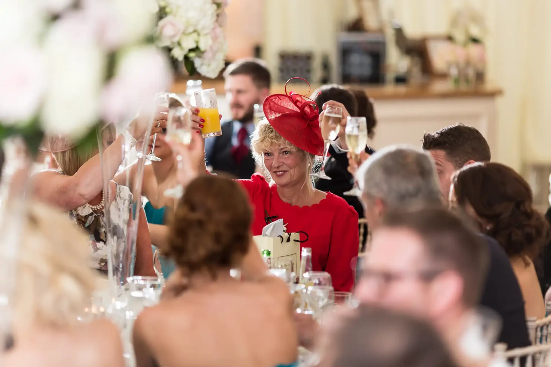 A woman in a red outfit and fascinator toasting with a drink at a crowded wedding reception.