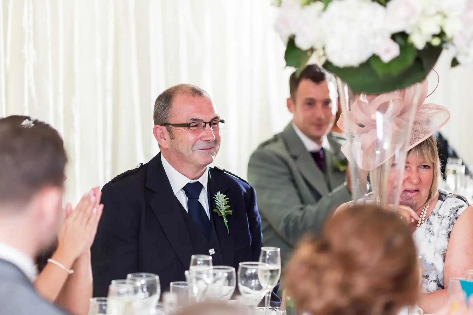 A man in a suit with a boutonniere smiles during a wedding reception, seated at a table with guests and floral decorations.