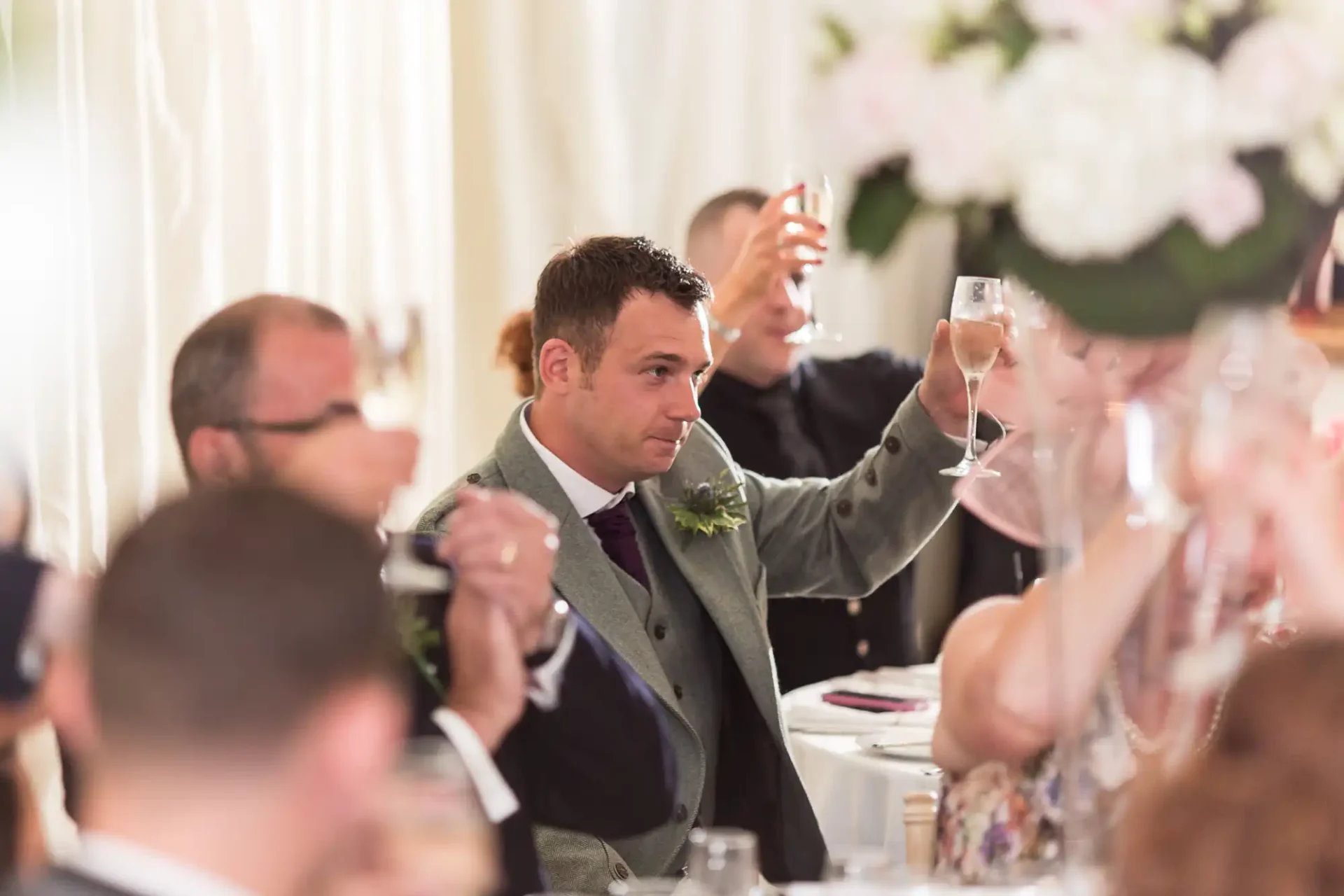 A man in a grey suit raising a toast with a glass of wine at a wedding reception, surrounded by guests.