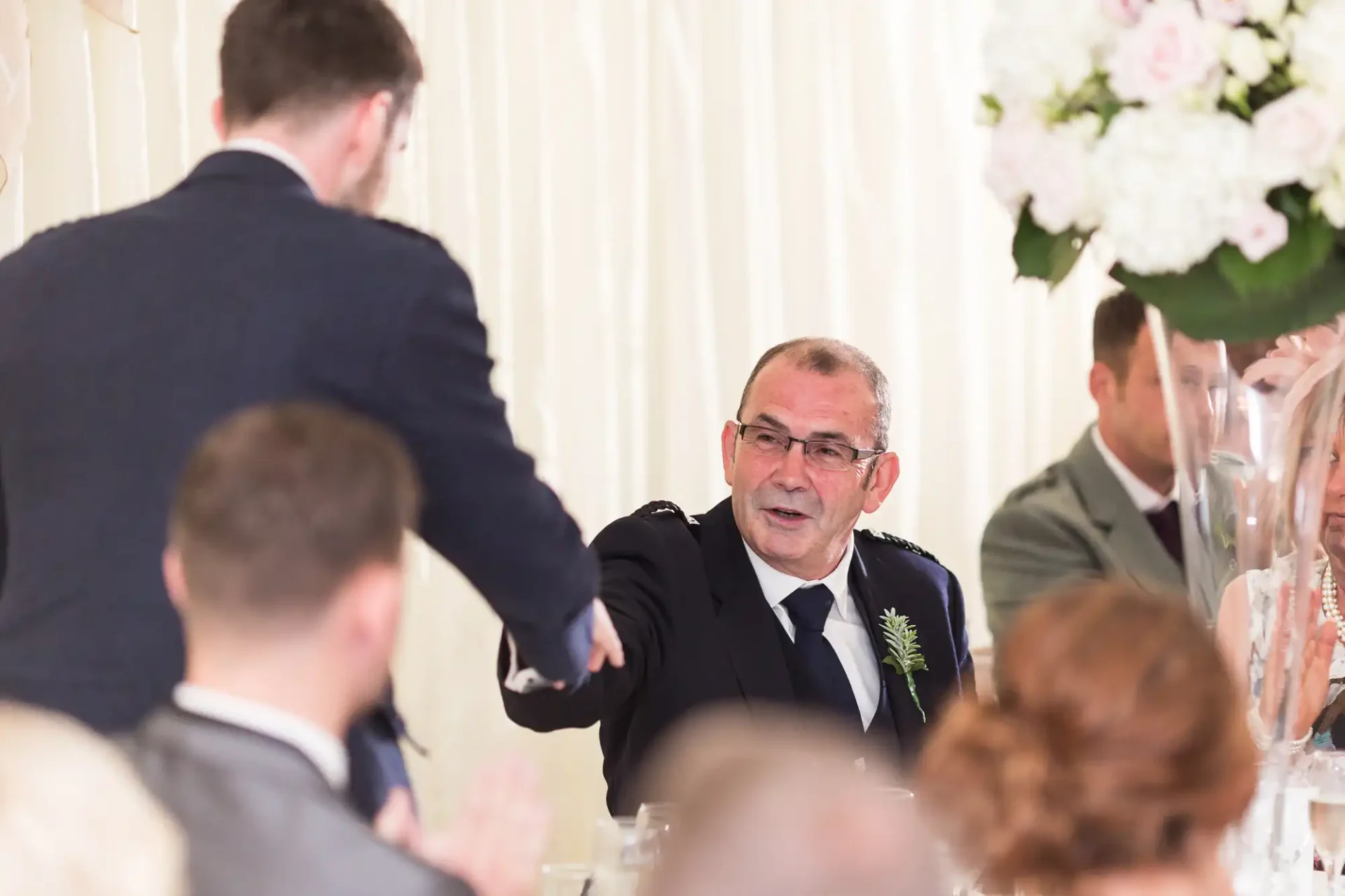 A man at a wedding reception extends his hand for a handshake, smiling, as guests around the table look on.