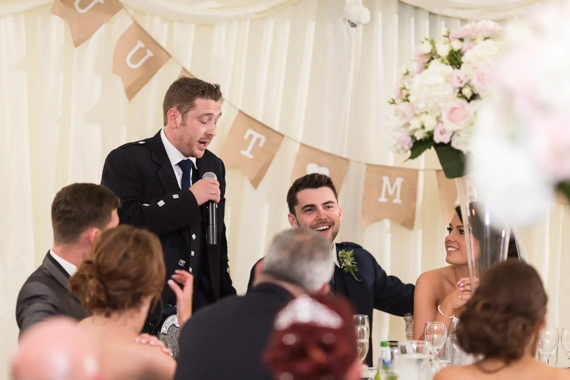 A man in a military uniform gives a speech at a wedding reception, holding a microphone, with guests listening and smiling.