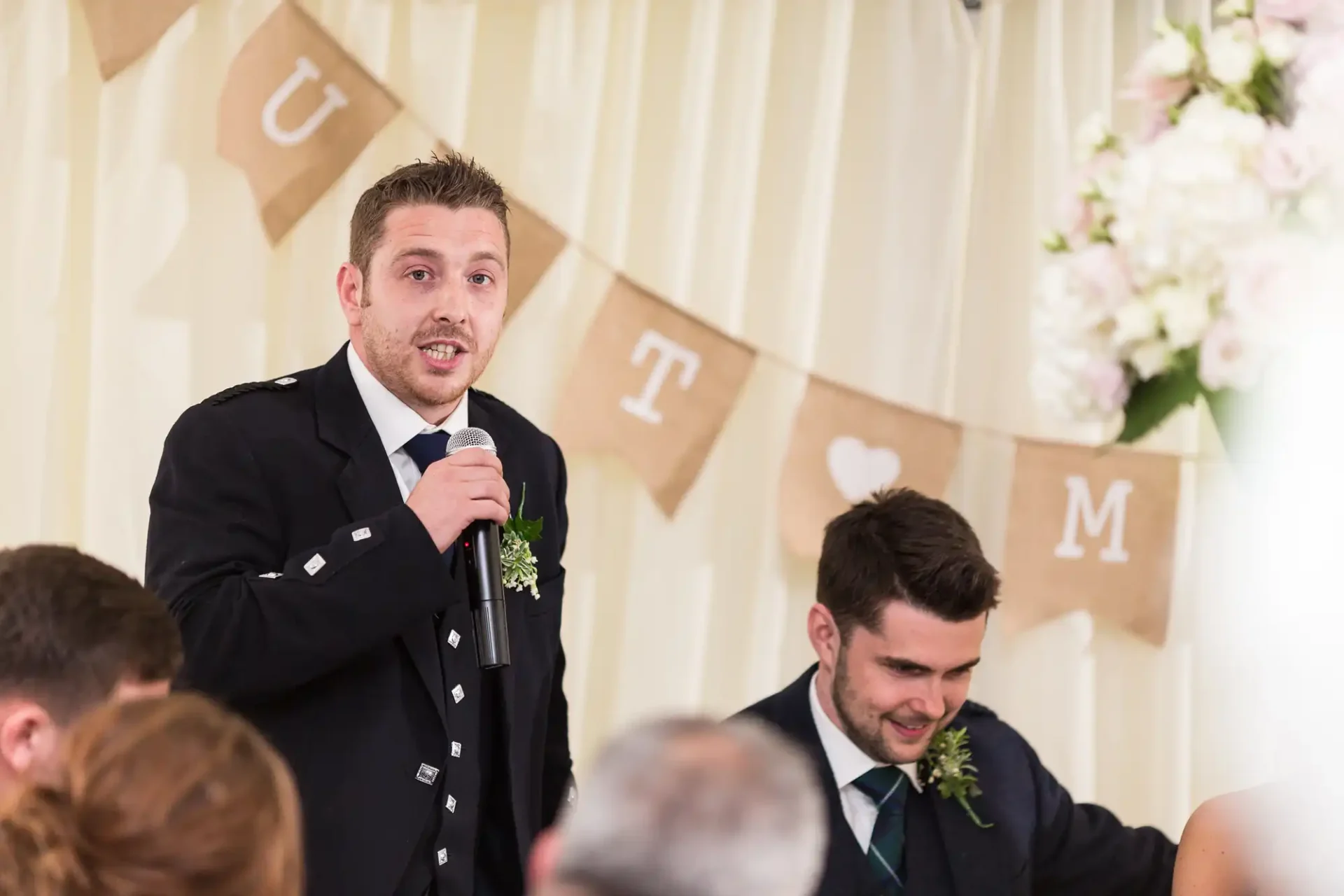 A man in a formal uniform giving a speech with a microphone at a wedding reception, another man smiling at him from a table.