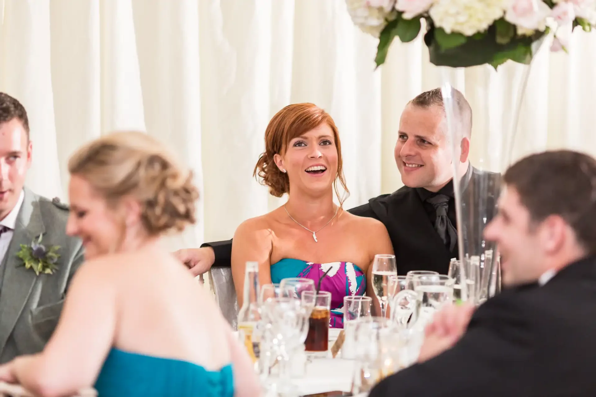 A joyful bride and groom sitting at a wedding reception table surrounded by guests, with floral decorations visible.