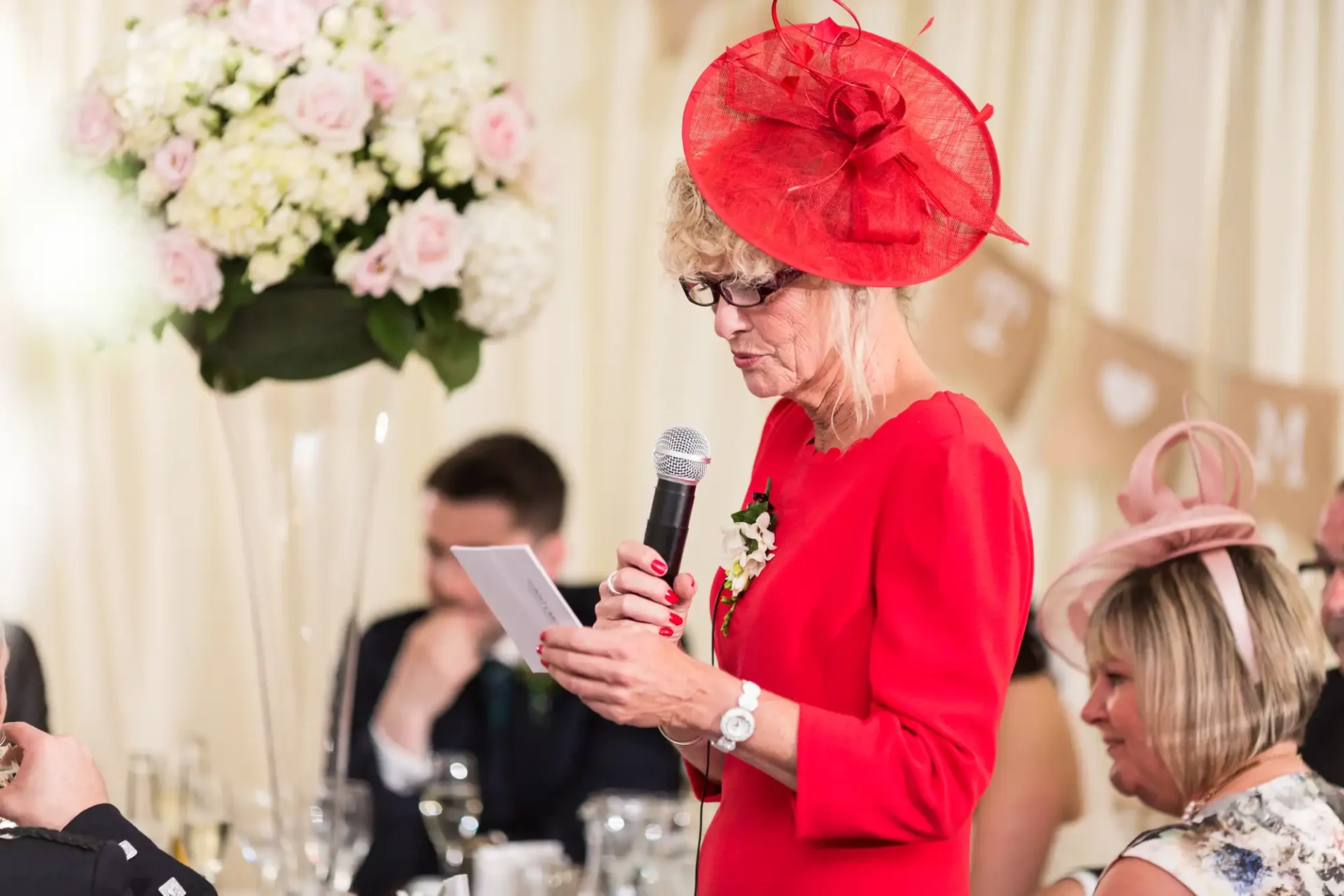 A woman in a red dress and hat speaks into a microphone while reading from a paper at a wedding reception.