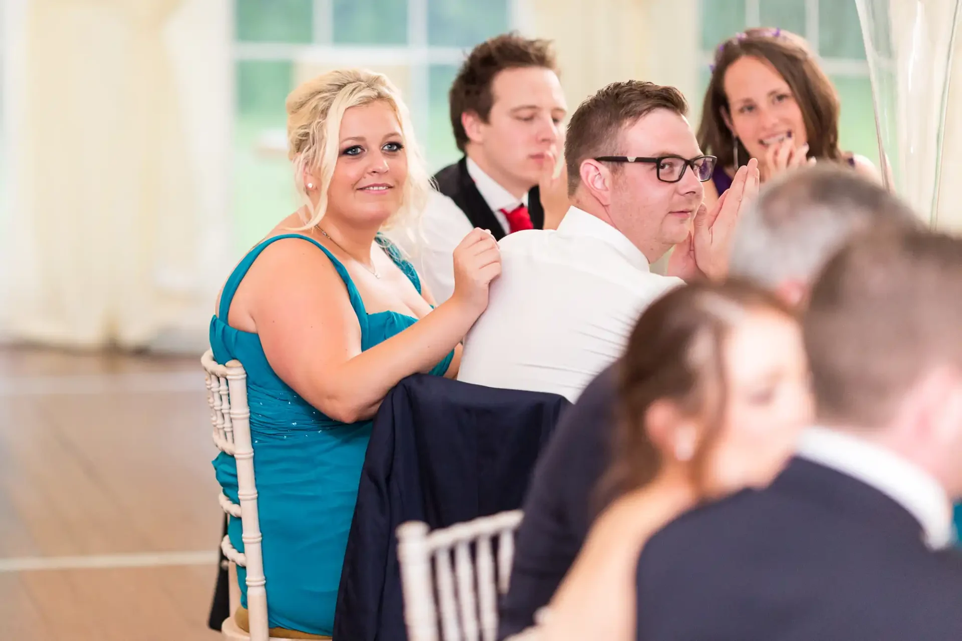 A group of guests at a wedding reception, focused on events off-camera, with one woman in a turquoise dress looking directly at the camera.