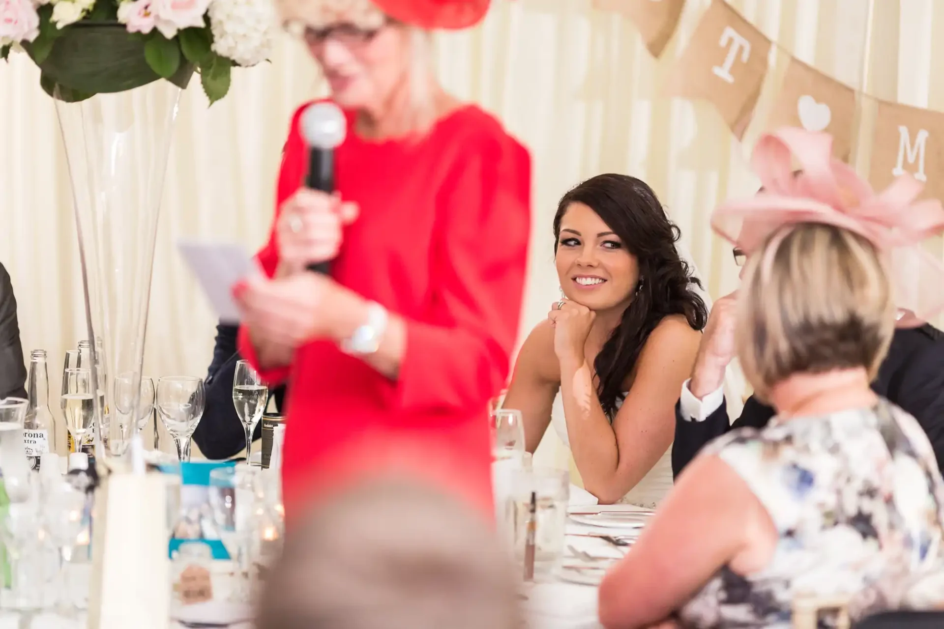 A woman in a red dress speaks into a microphone at a wedding reception, while a smiling young woman in the background looks on.