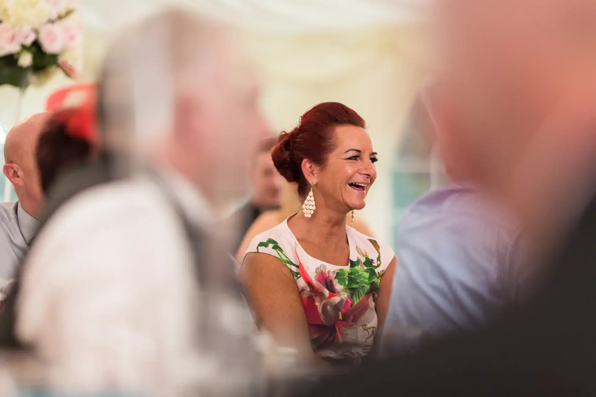 Woman with red hair laughing at a table during an event, surrounded by guests in soft focus.