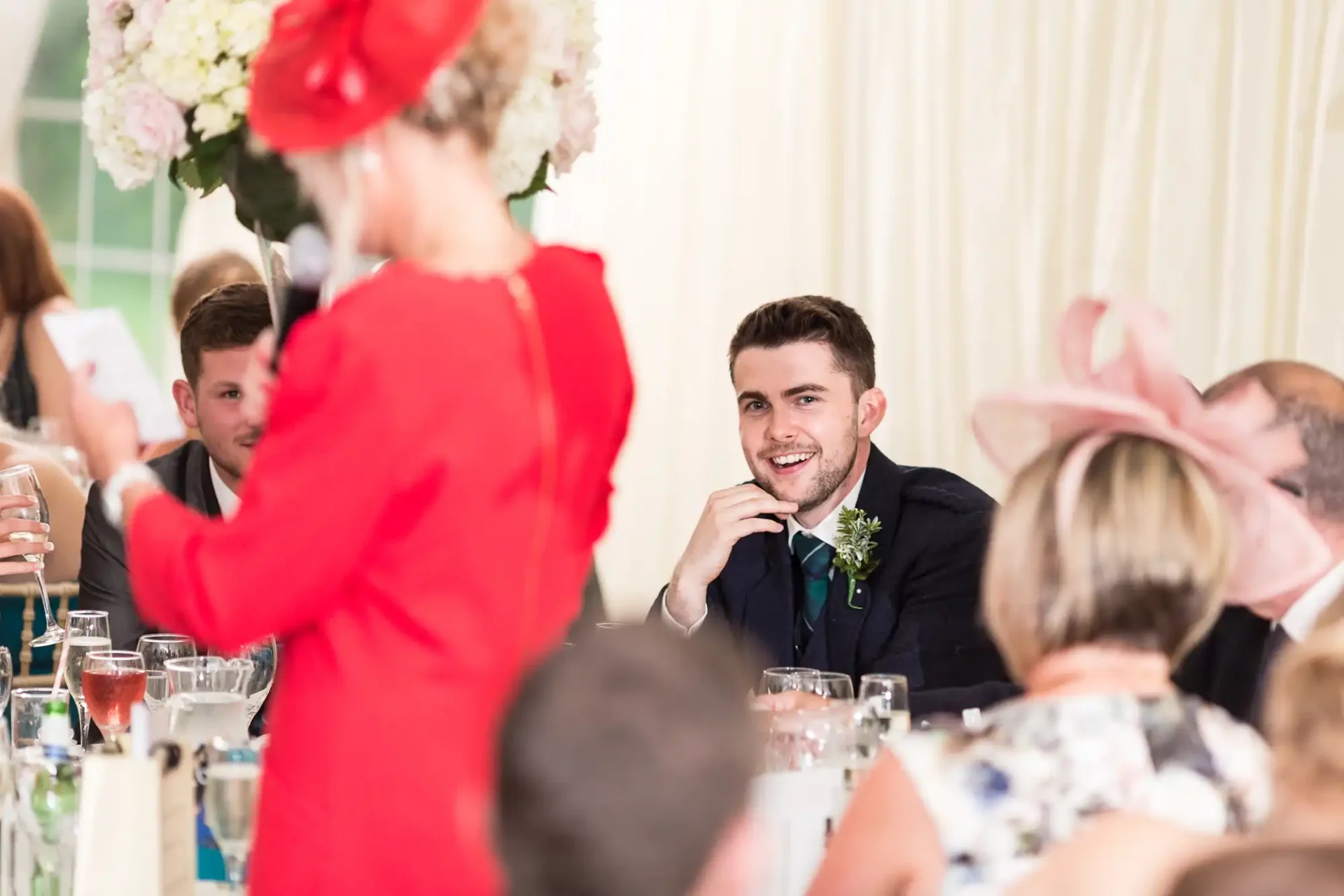 A man in a suit with a boutonniere smiling at a woman in a red dress at a wedding reception, with guests blurred in the background.