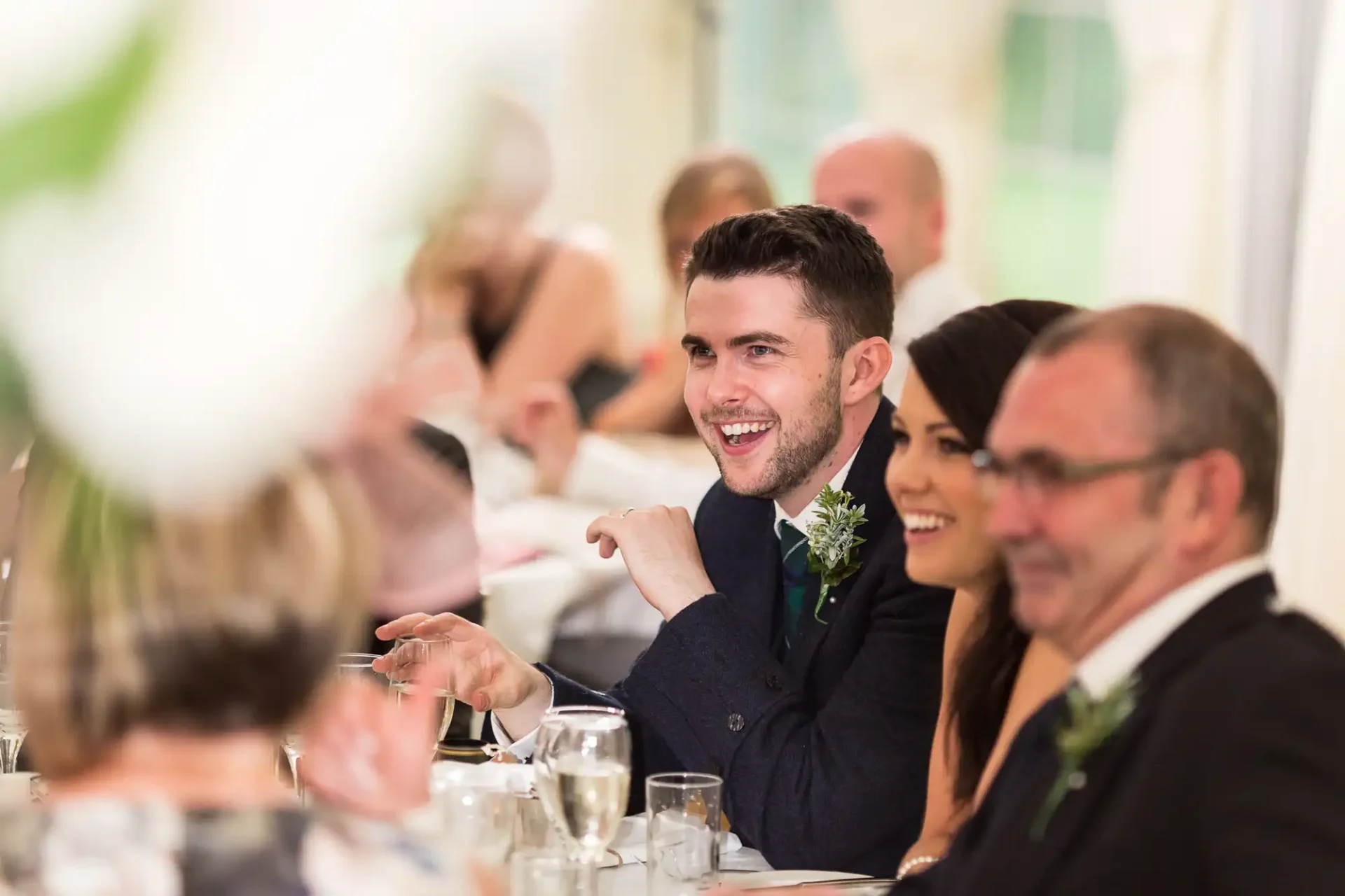 A groom sitting at a wedding reception table, smiling and conversing with guests, with a focus on his joyful expression.
