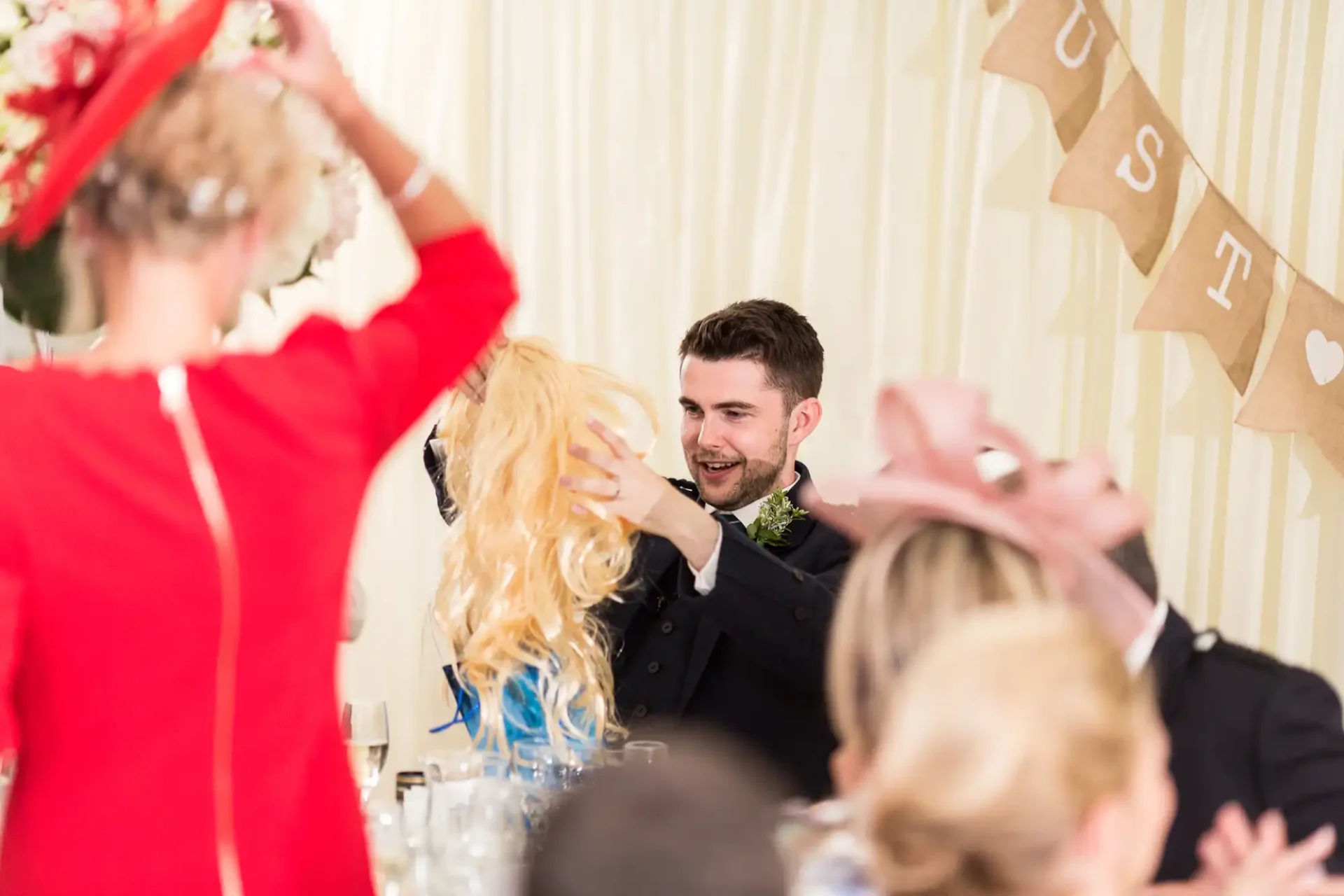 A groom smiles while engaging in a lively wig-toss game with guests at a wedding reception, under a banner that reads "just married.