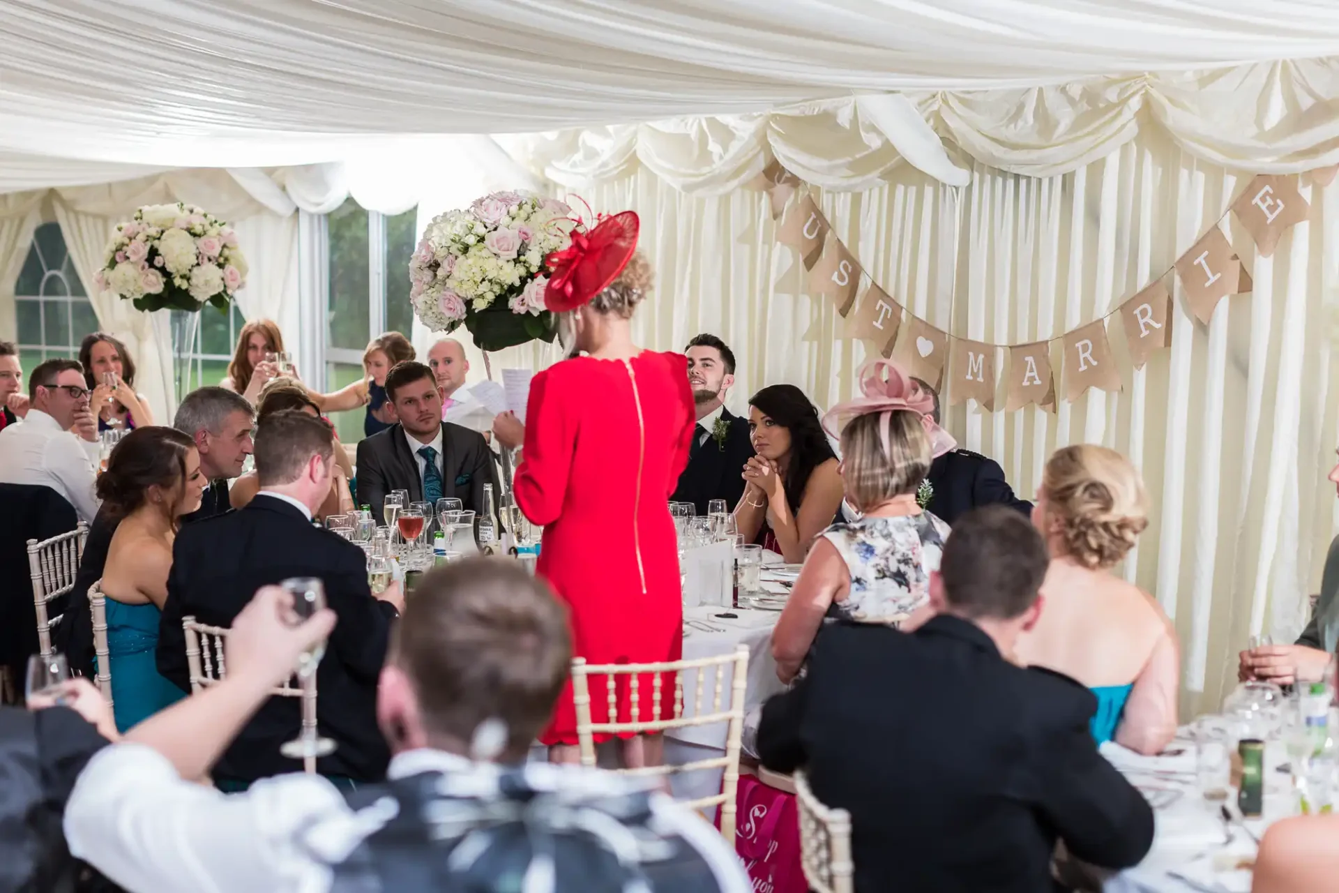A woman in a red hat makes a speech at a wedding reception inside a tent adorned with "just married" banners, as guests listen attentively.