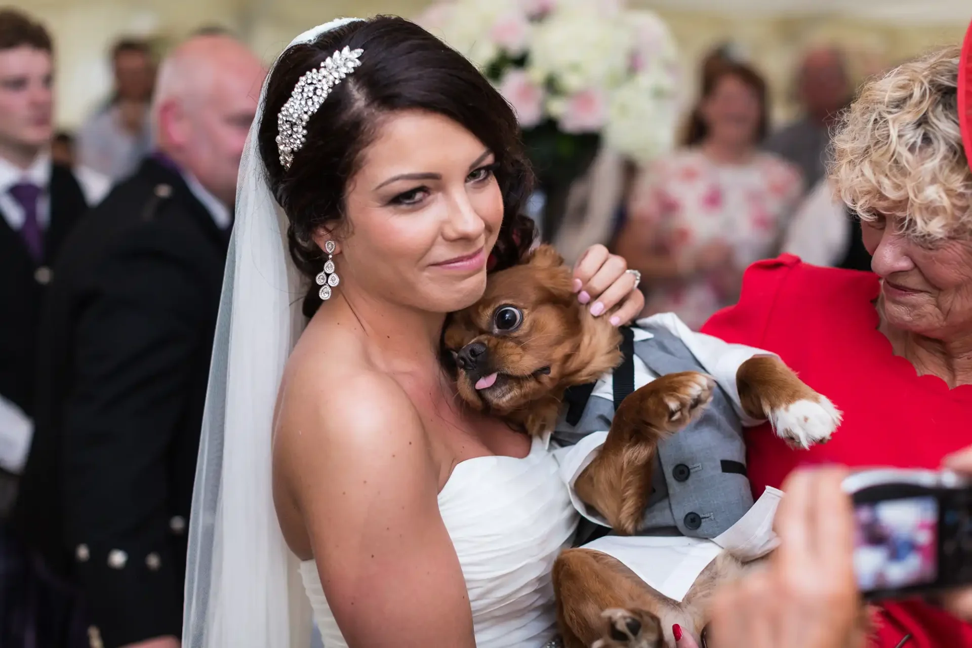 A bride holding a small brown dog smiles during a wedding reception as an elderly woman assists and guests watch.