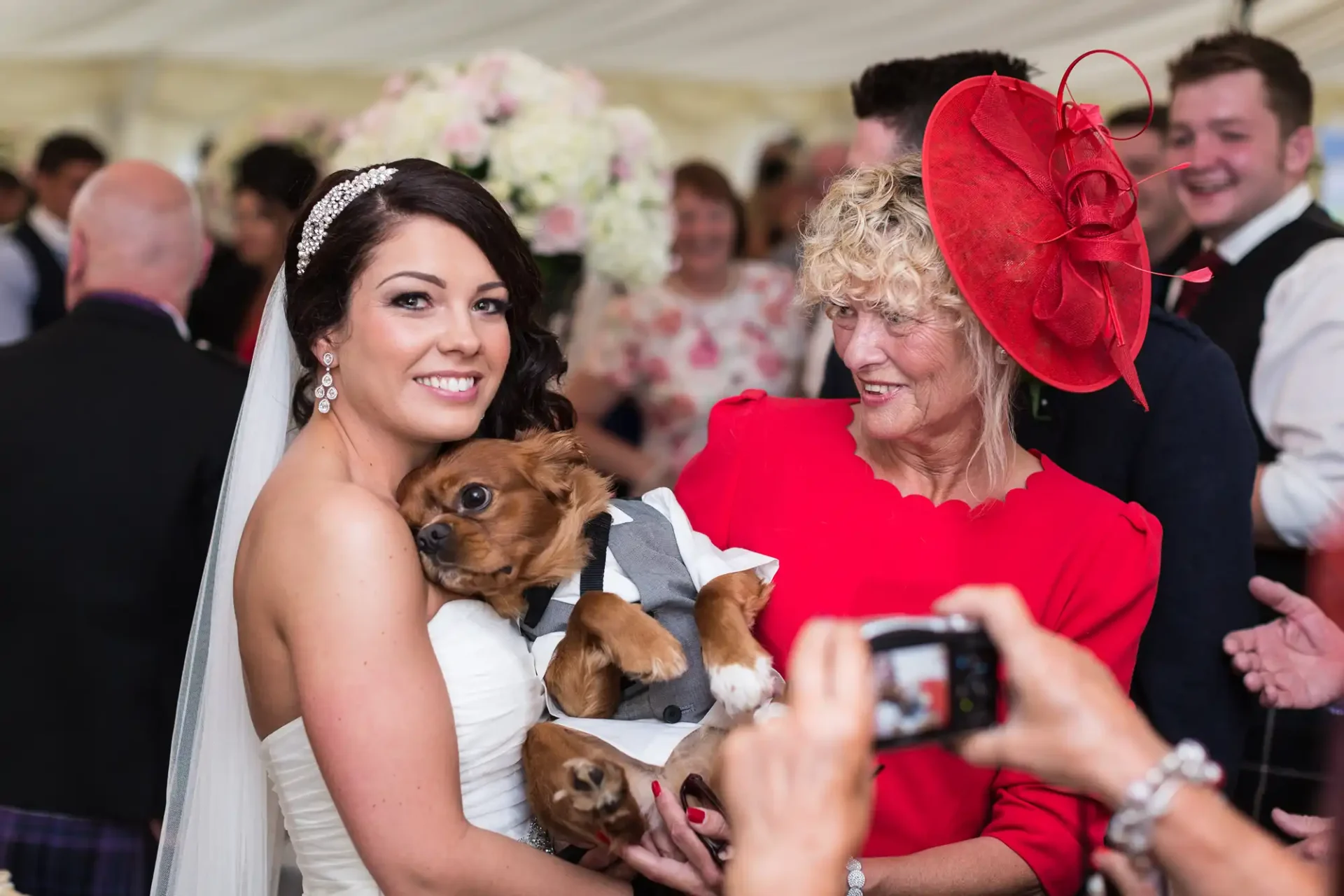 A bride in a white dress holding a small dog, with an older woman in a red outfit and hat smiling at her, in a crowded event tent.
