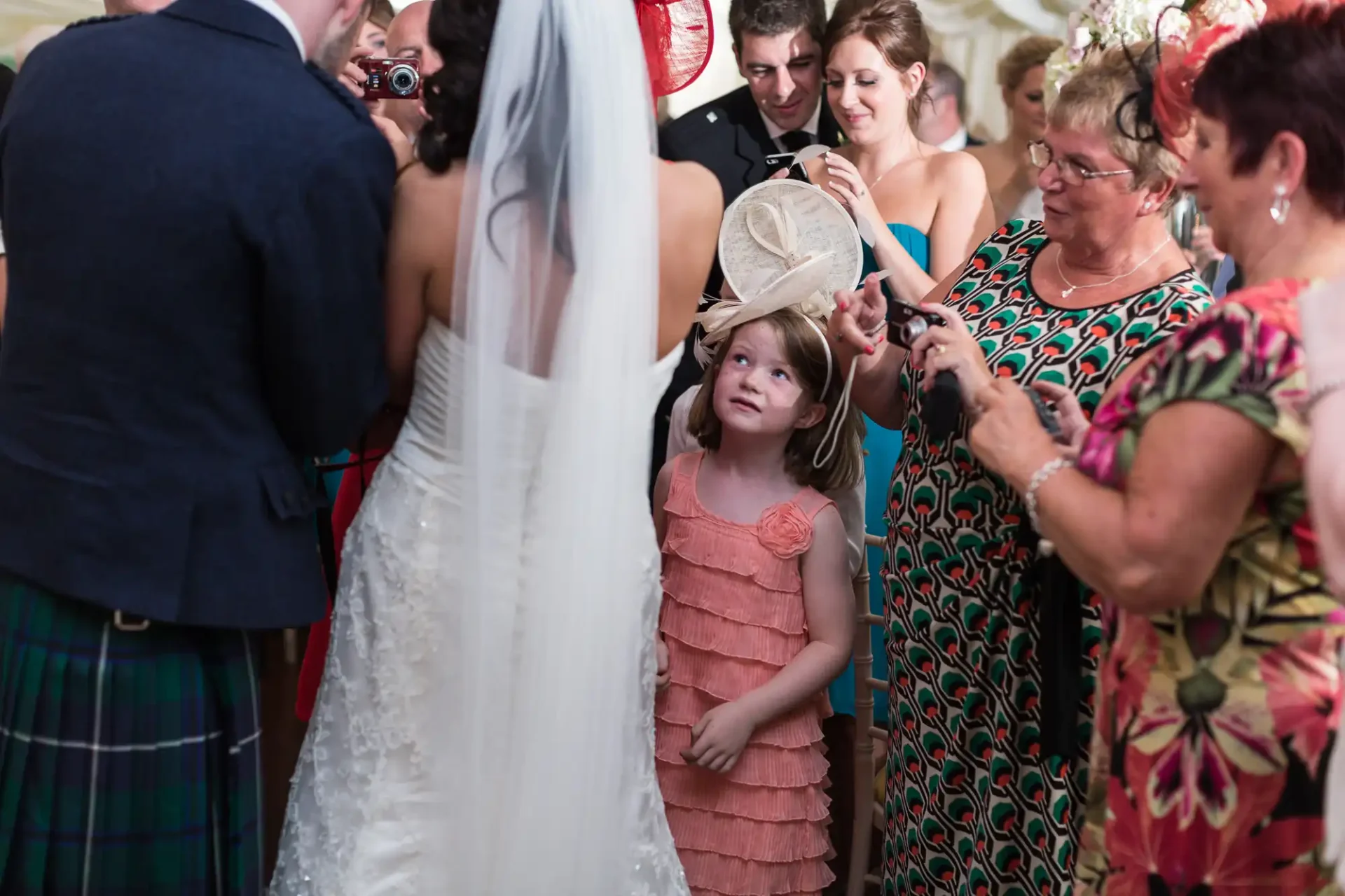 A young girl in an orange dress looking up curiously at a bride in a white gown surrounded by guests at a wedding.