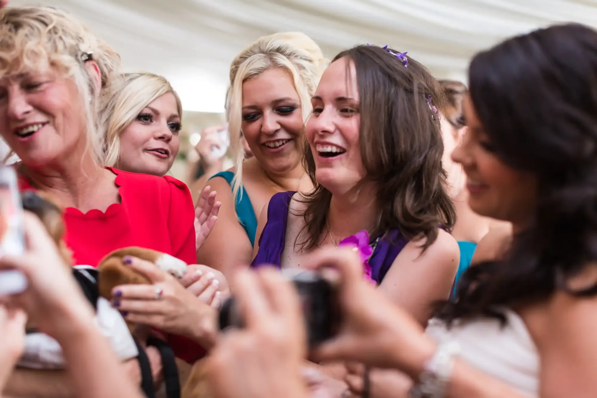 Group of women laughing and clapping at a social event under a tent, one capturing the moment on her smartphone.