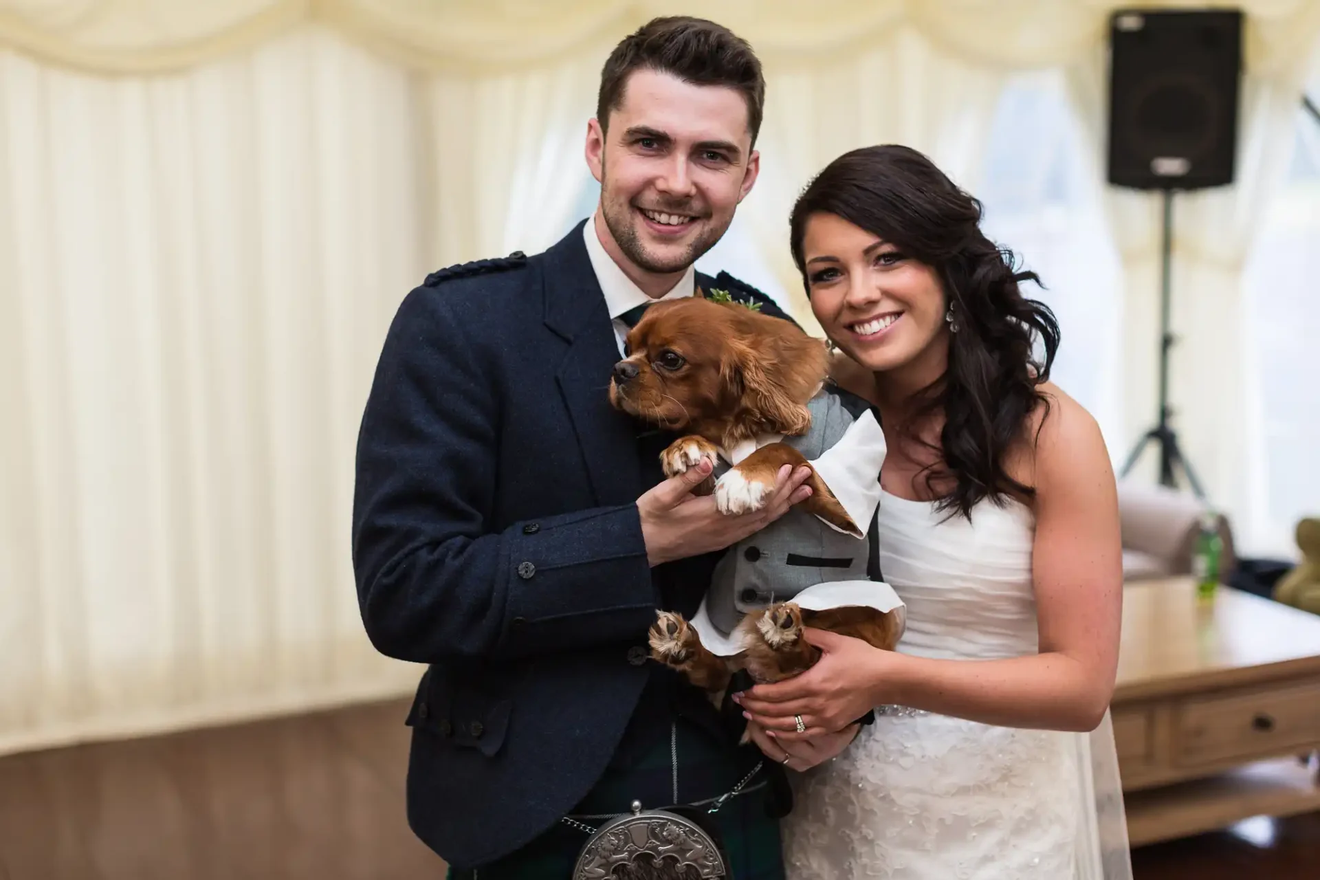 A smiling bride and groom, dressed in formal wedding attire, hold a small brown dog at their wedding reception.