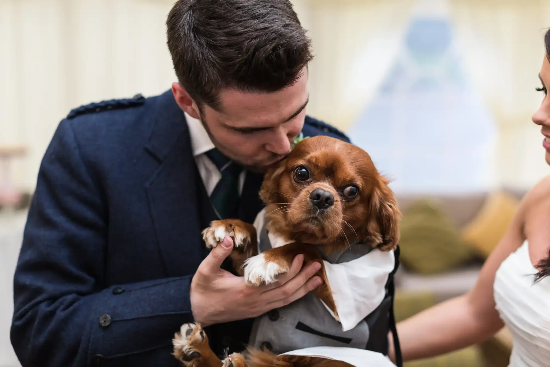 Groom in a suit kisses a brown and white cavalier king charles spaniel held by a bride in a white dress.