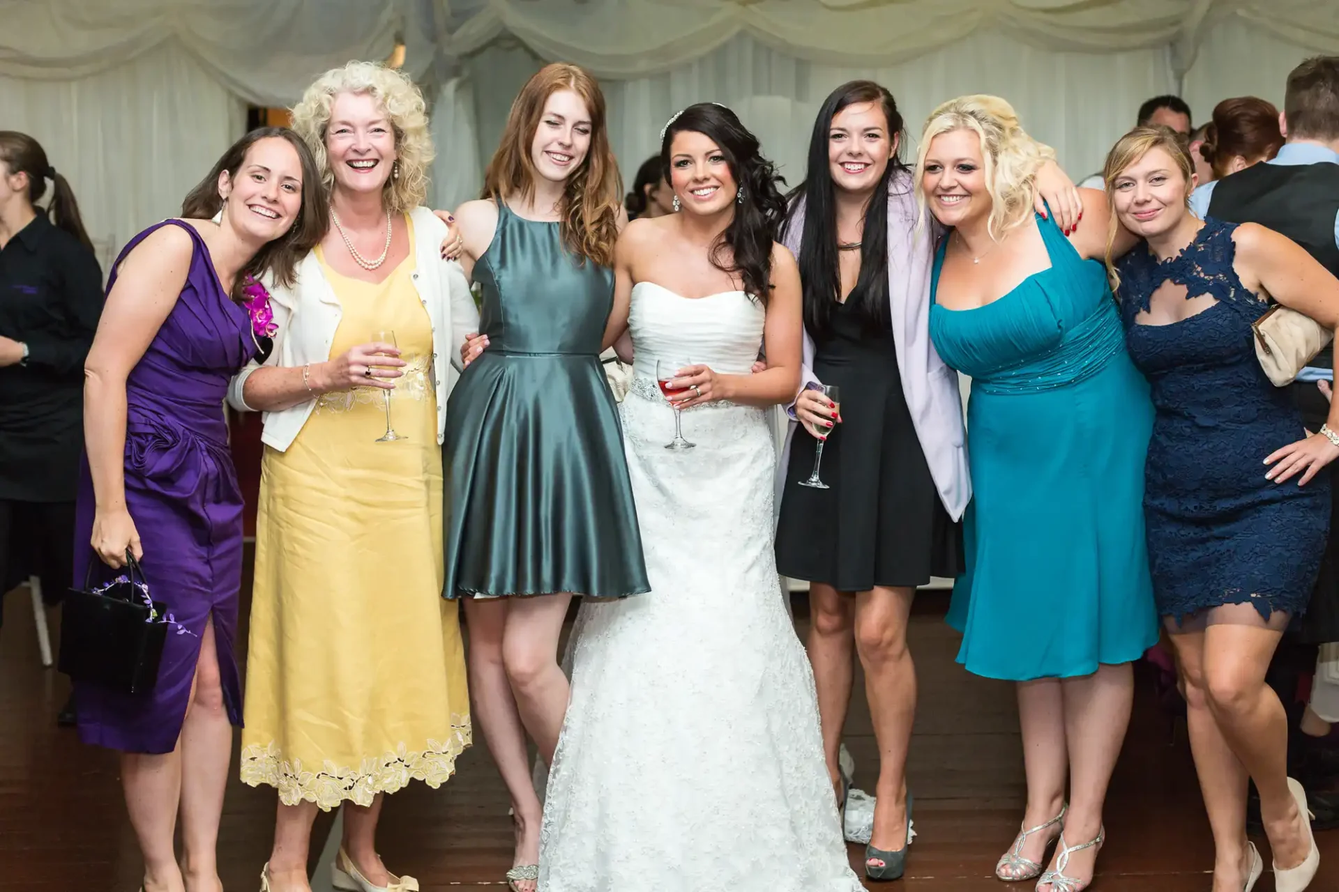 Six women posing for a photo at a wedding reception, smiling and holding champagne glasses, with one in a bridal gown.