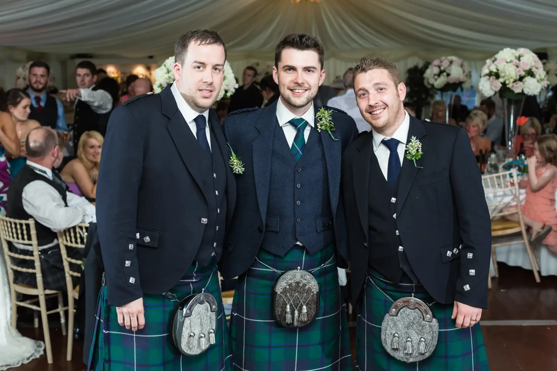 Three men in traditional scottish kilts with sporran, smiling and posing together at a wedding reception inside a tent.