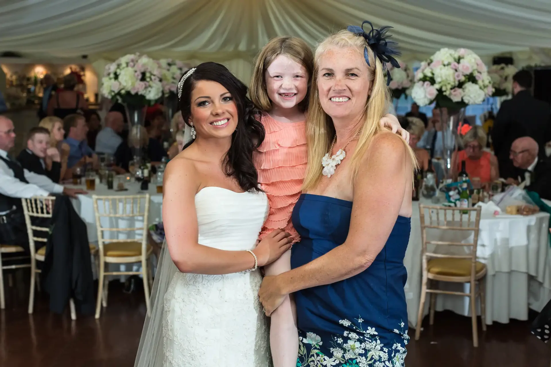 Three generations of women, a bride in white, a young girl, and an older woman in blue, smiling together at a wedding reception.