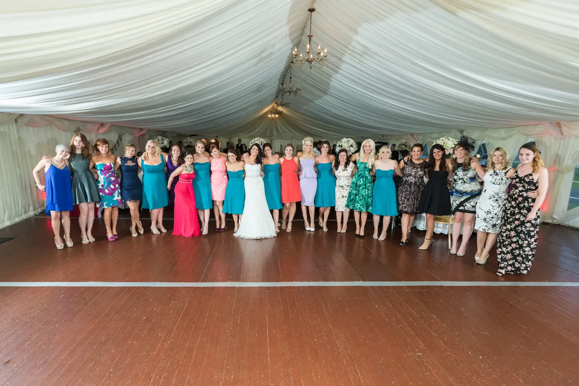 A group of women in formal dresses posing together at a wedding reception inside a tent with draped ceilings and a chandelier.